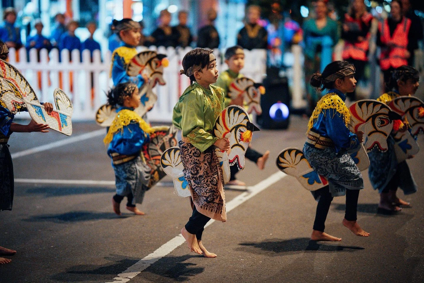 Children participating in a traditional cultural dance parade at night, dressed in colorful costumes, carrying decorative fans, walking barefoot on the street.