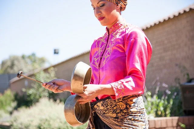 Woman in traditional Indonesian attire holding brass gongs outdoors.