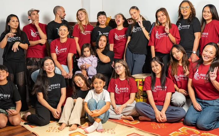 A group of about 16 people in red or black Suara t-shirts pose. Kids are sitting at the front.