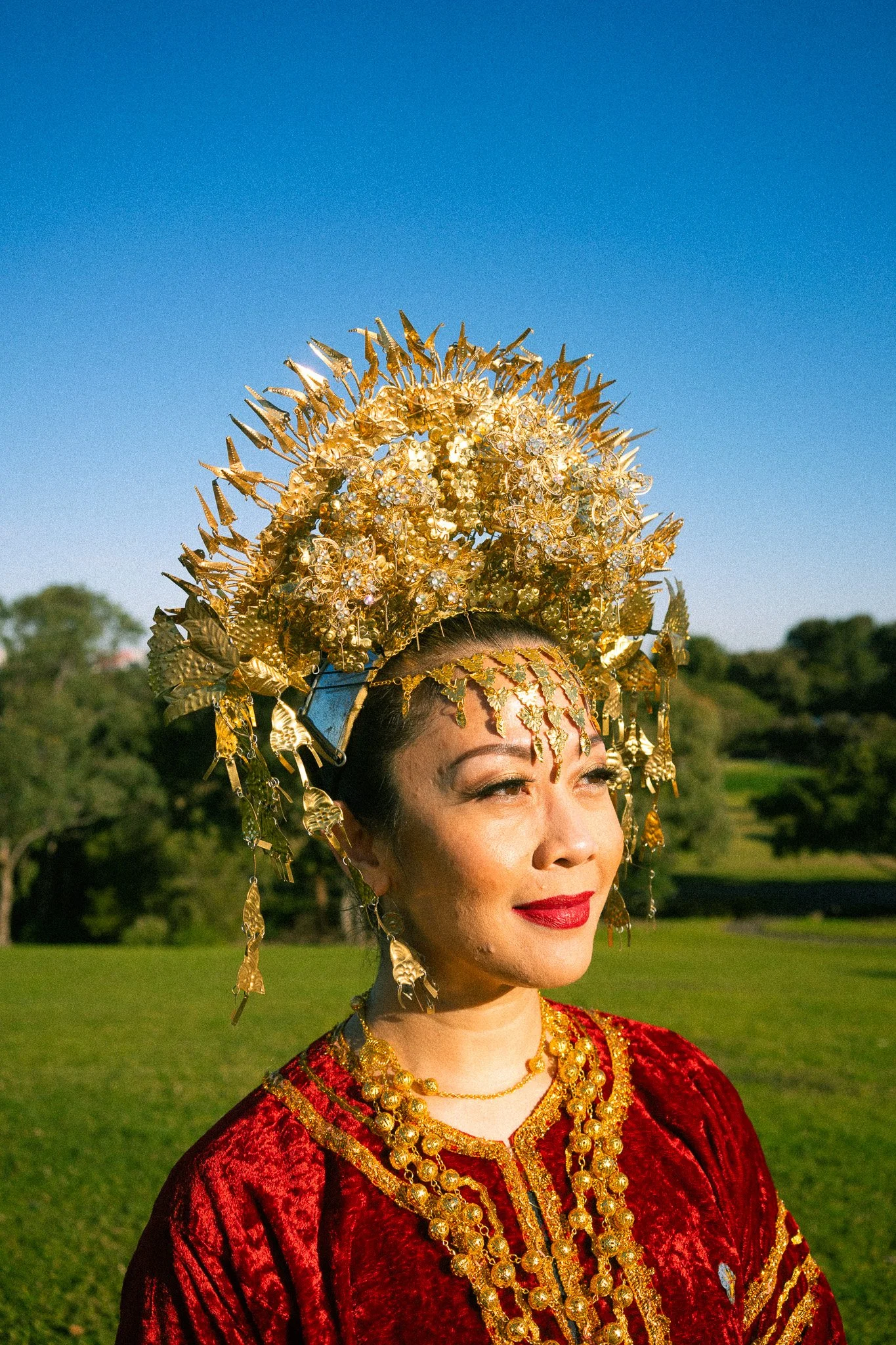 A woman in traditional attire with a large, ornate gold headdress and matching jewelry, standing outdoors on a sunny day with a clear blue sky and green trees in the background.