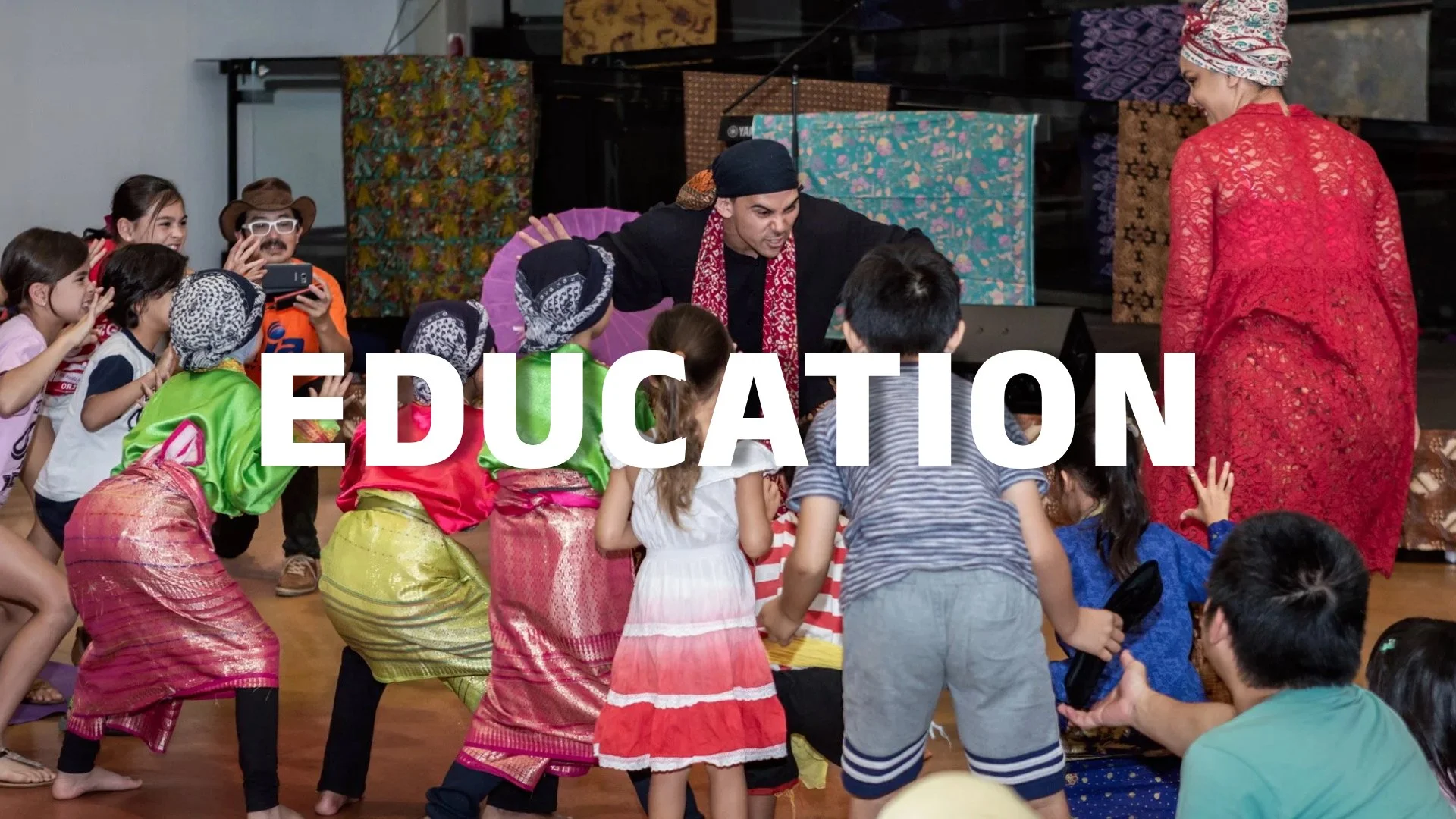 Children in colorful traditional clothing participating in a cultural dance or activity with an adult, indoors, with the word 'EDUCATION' overlayed.
