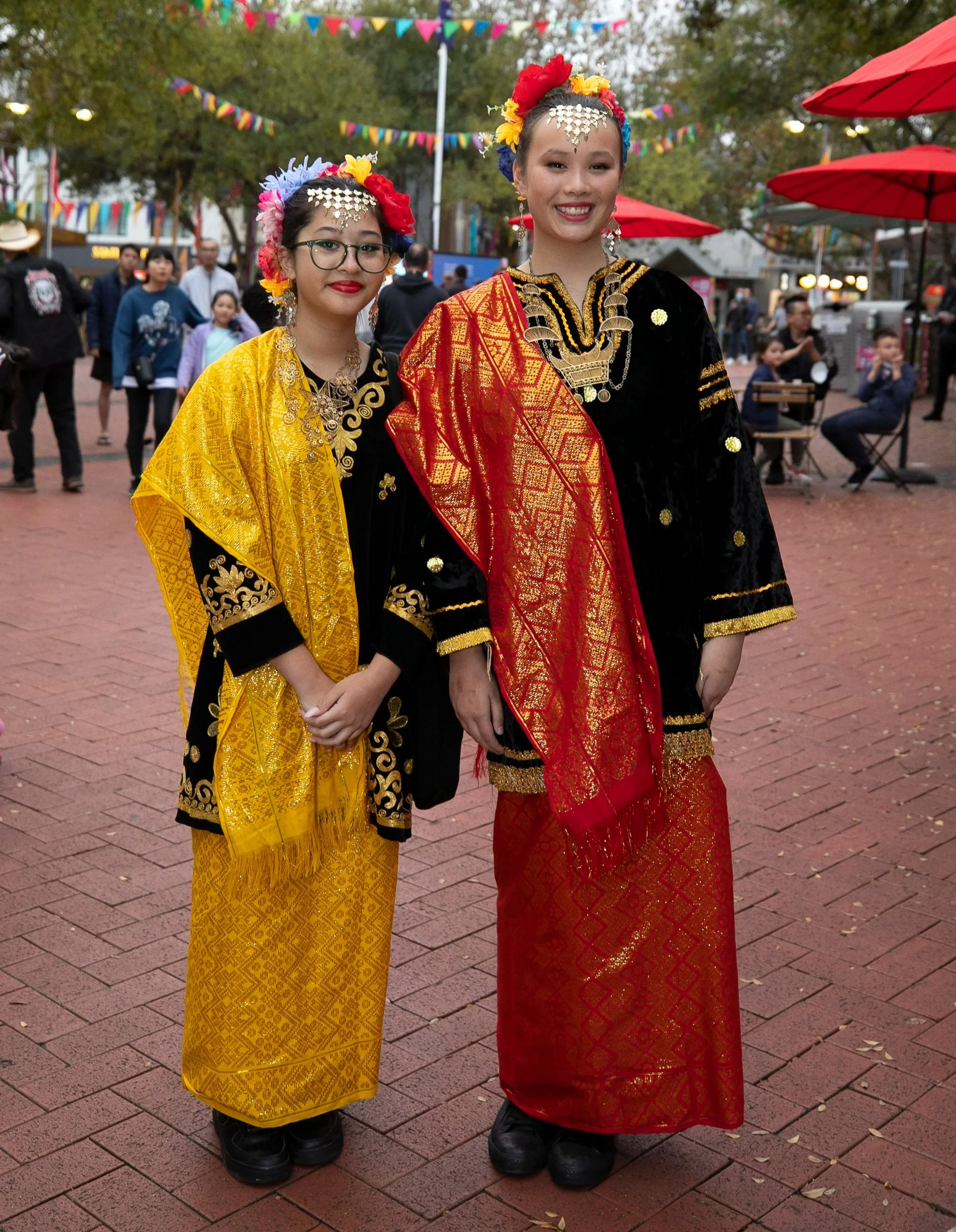 Two women dressed in traditional Indonesian attire with vibrant yellow and red shawls, standing outdoors on a red brick pavement during a festival or celebration, with colorful banners and other people in the background.