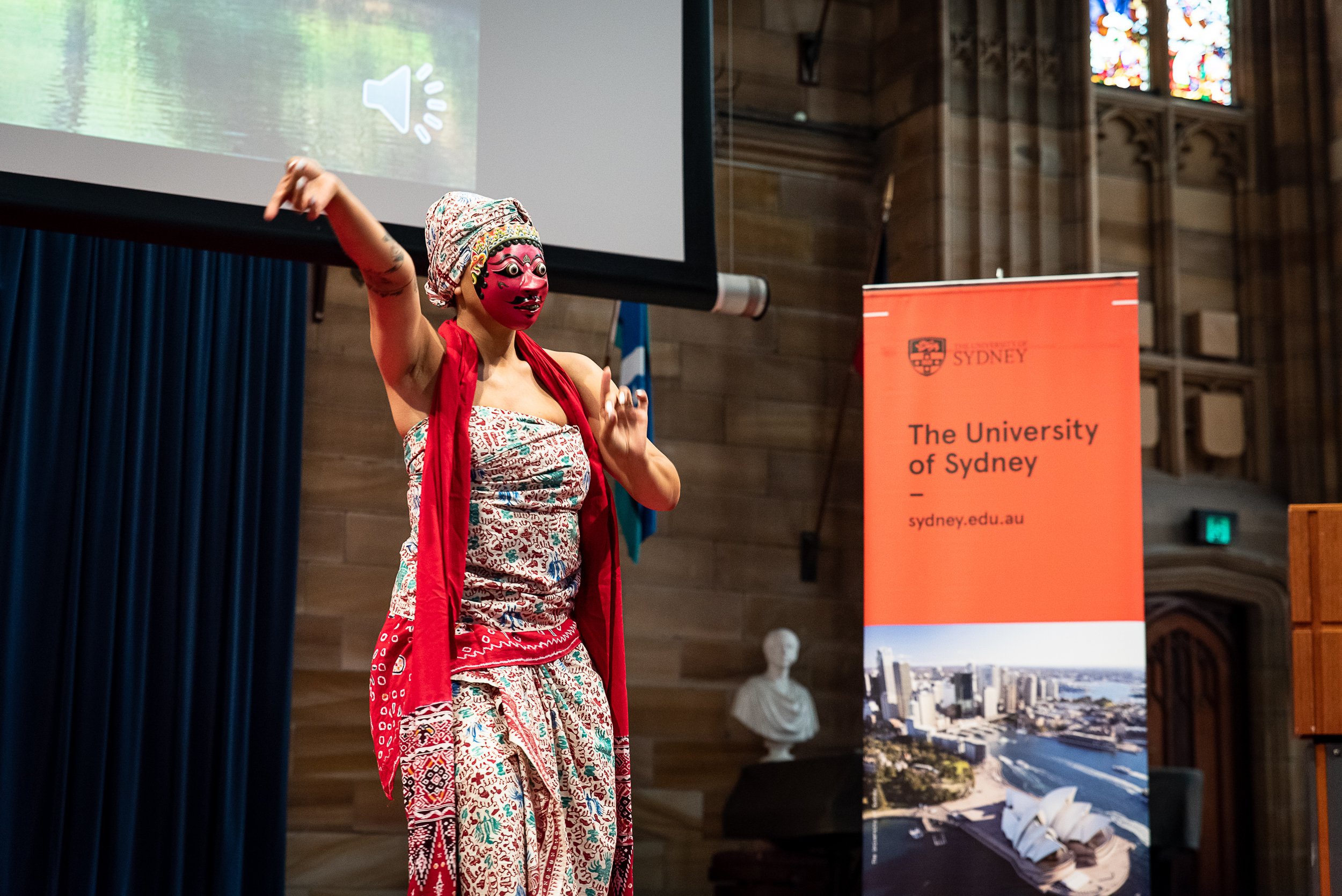 Person wearing a traditional mask and colorful clothing, speaking or performing at an event at the University of Sydney, with a banner displaying the university's logo and a cityscape.