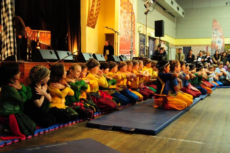 Children performing a mass seated dance in traditional colorful clothing, seated cross-legged on mats, with an audience in the background.