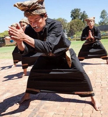 A group of men dressed in traditional Indonesian black clothing practicing a martial arts or dance move outdoors on brick pavement.