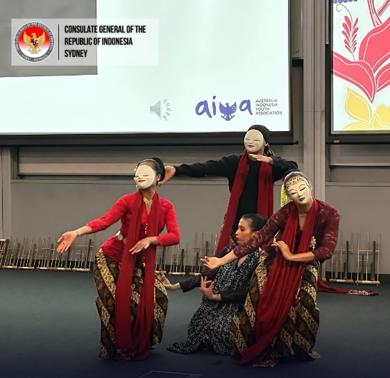 Four performers in traditional Indonesian costumes and masks performing a dance on stage, with a large screen behind displaying logos and text.