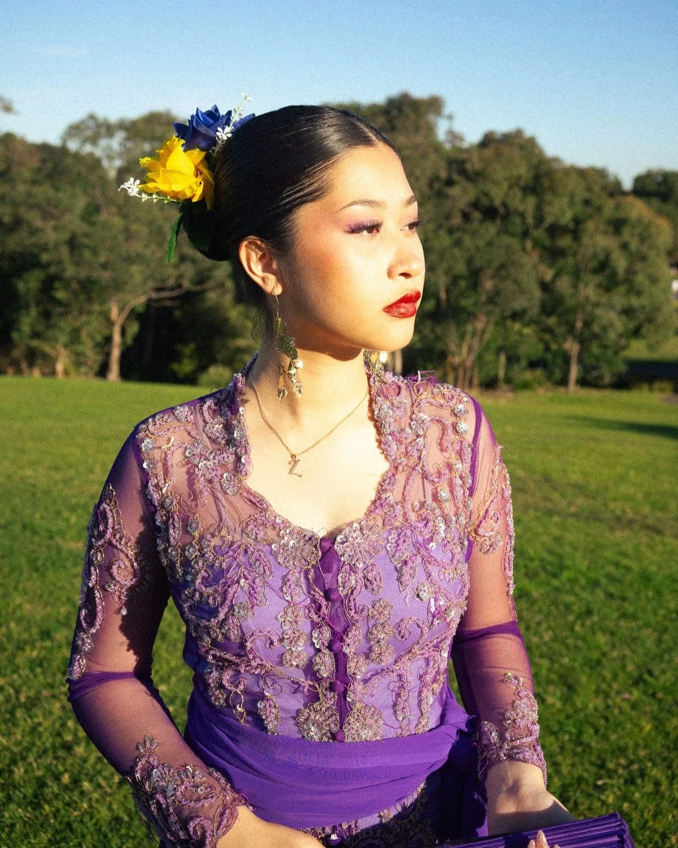 A woman wearing a purple embroidered dress with flowers and jewelry, standing outdoors in a grassy area with trees in the background, during daytime.