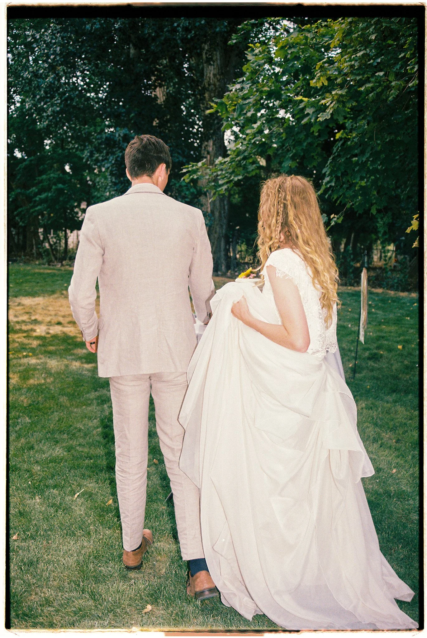 A bride and groom walking outdoors, the bride carrying her wedding dress, surrounded by green trees and grass.