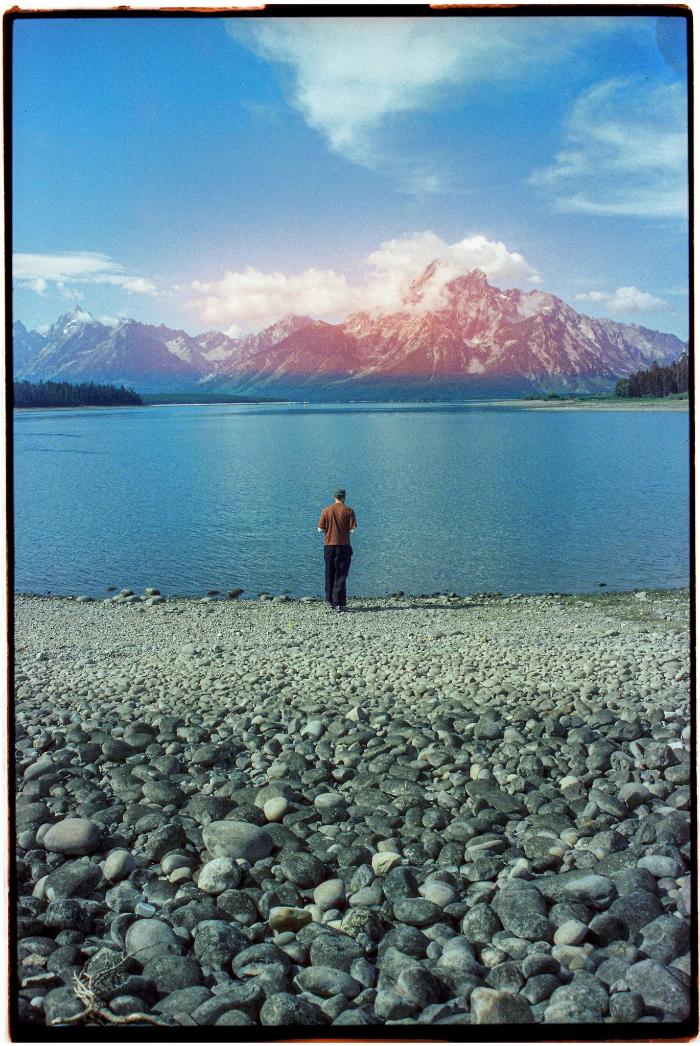 A person standing on a rocky shoreline facing a lake with mountains in the background and a partly cloudy sky.