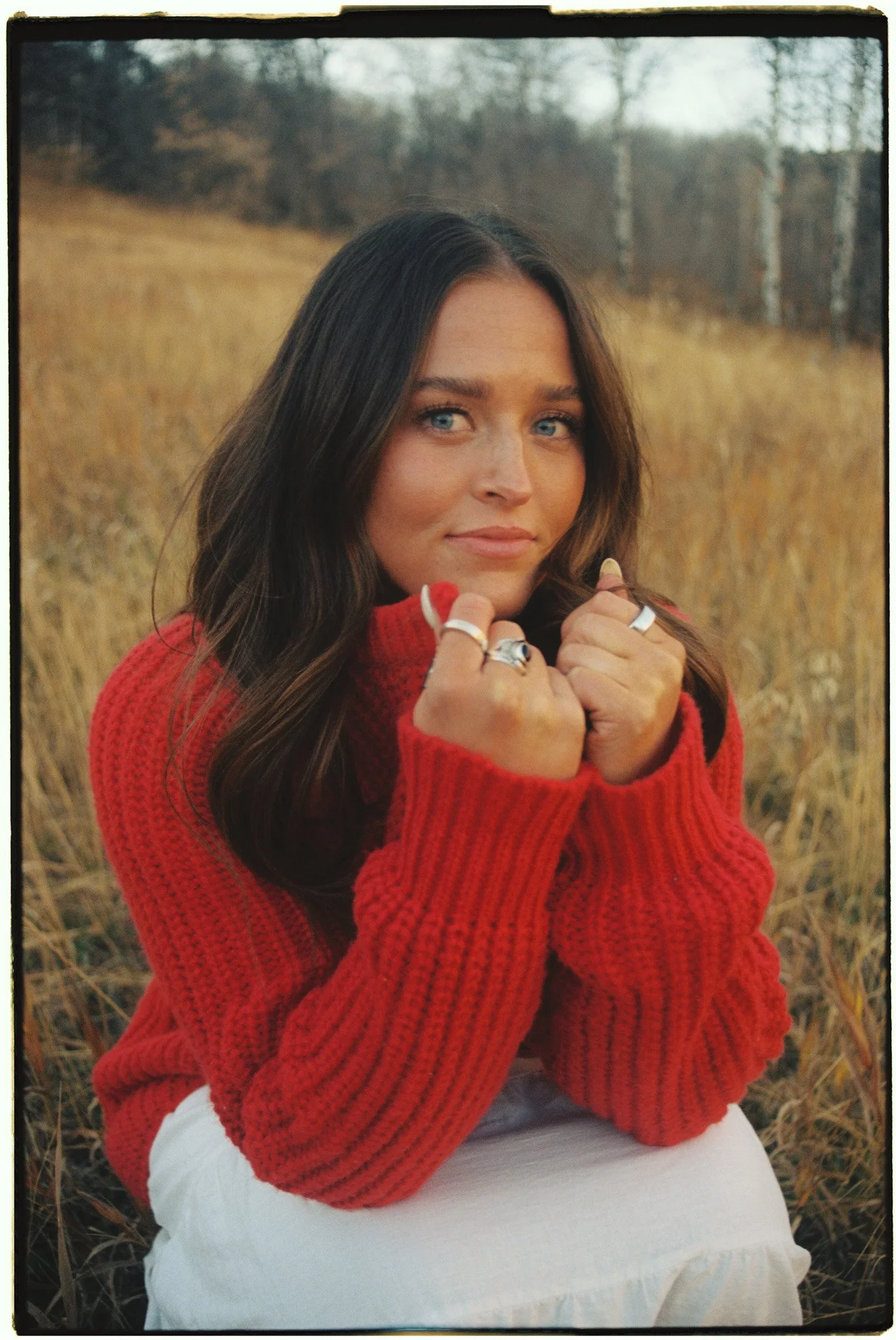 A young woman with blue eyes and brown hair, wearing a red knit sweater, sitting in a field of tall, dry grass with trees in the background during autumn.