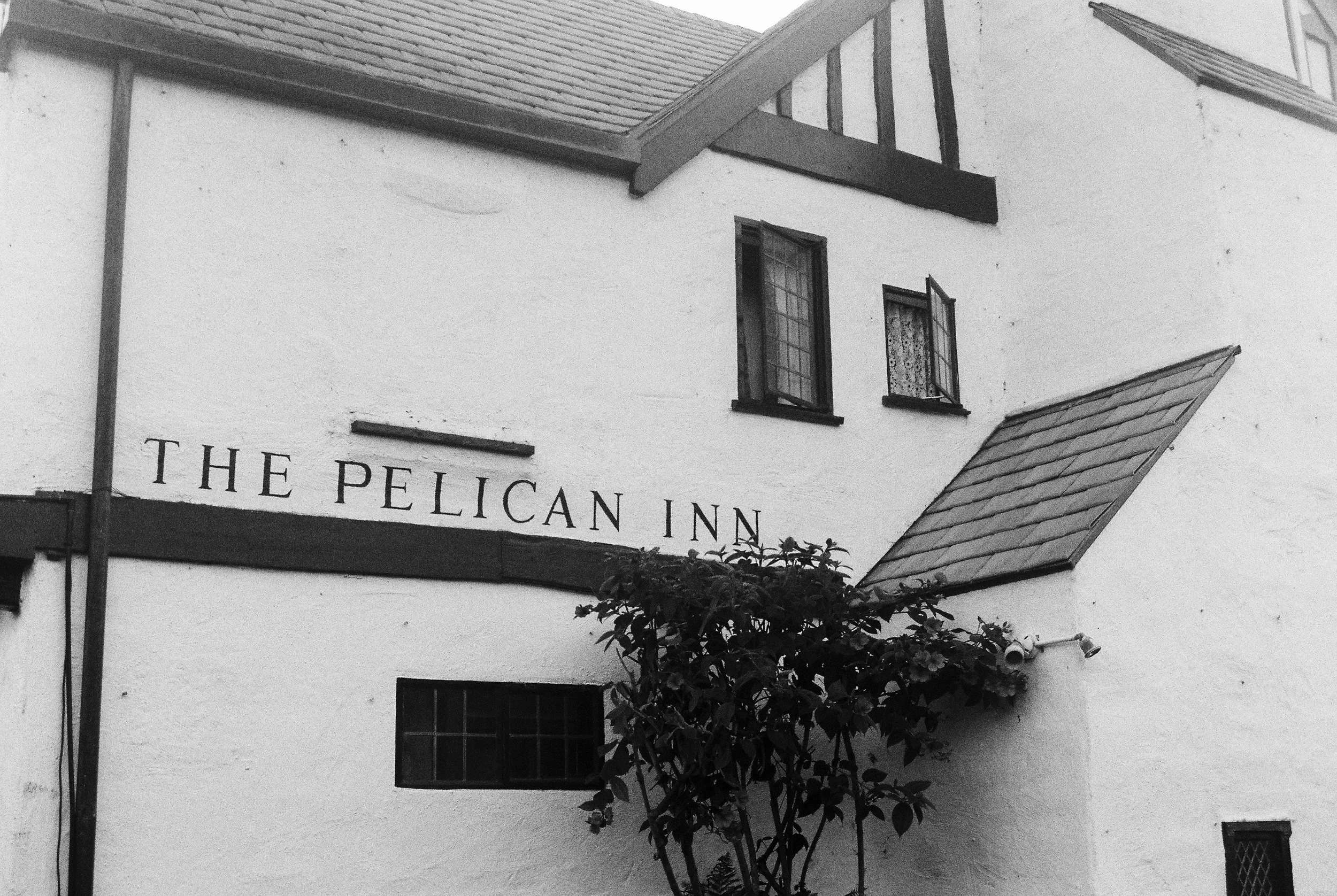 Exterior of The Pelican Inn, a building with white walls, small windows, and a sign with the name on it. There is a plant in front and a small security camera on the wall.
