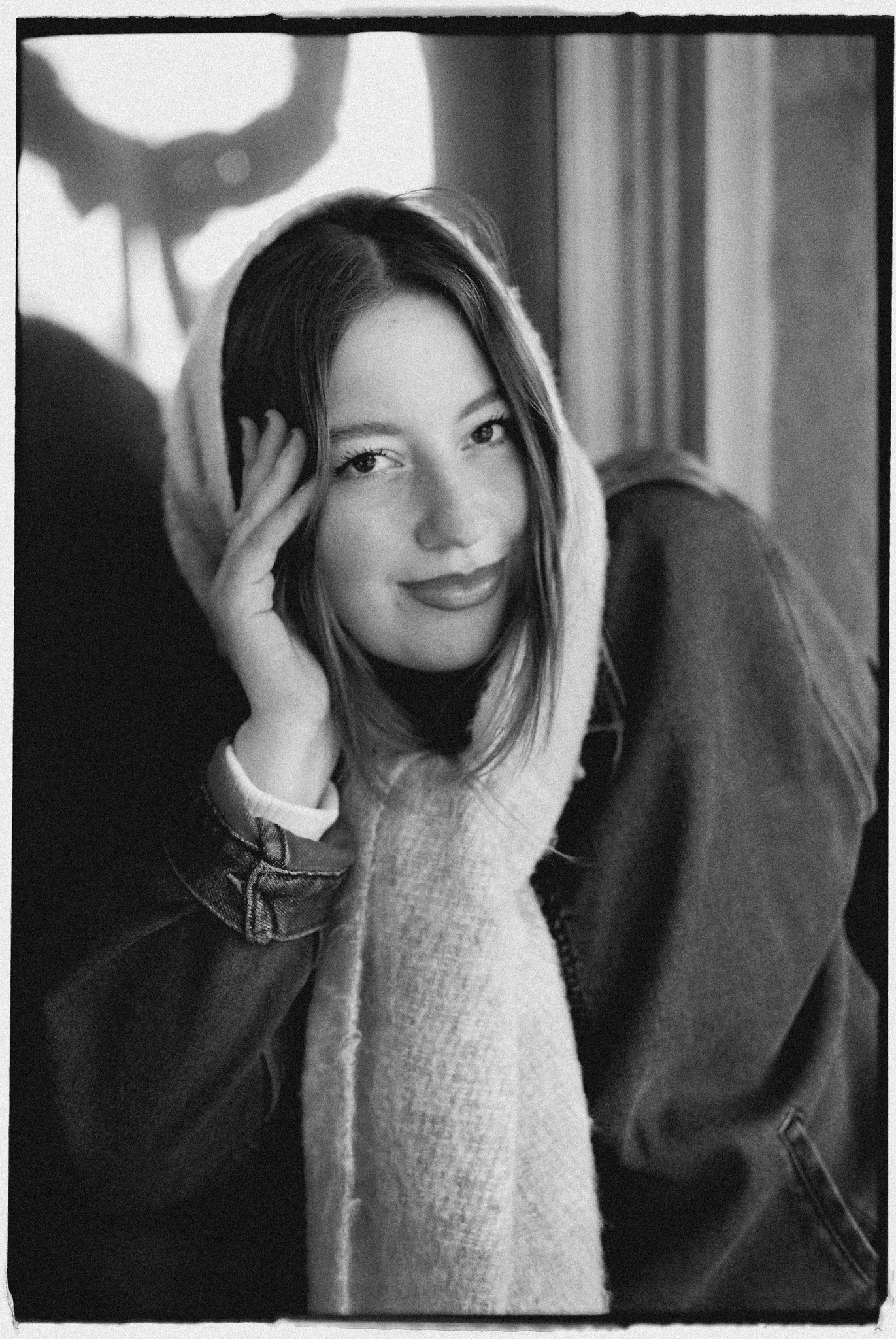 Black and white photo of a young woman with shoulder-length hair, smiling, resting her head on her hand, wearing a hooded sweatshirt and a denim jacket, sitting indoors near a window.