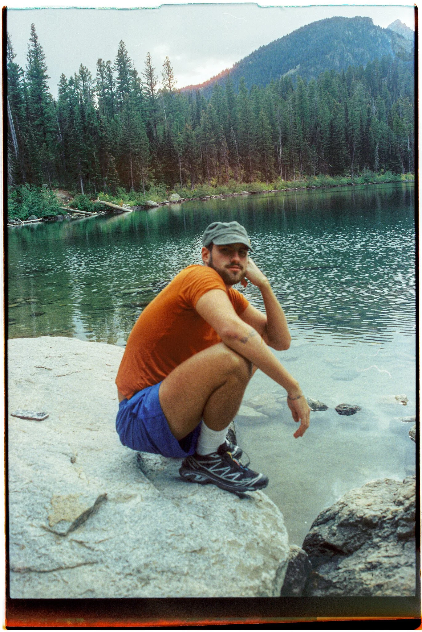 A man in an orange t-shirt, blue shorts, and a gray cap sitting on a rock by a lake with a forest and mountains in the background.