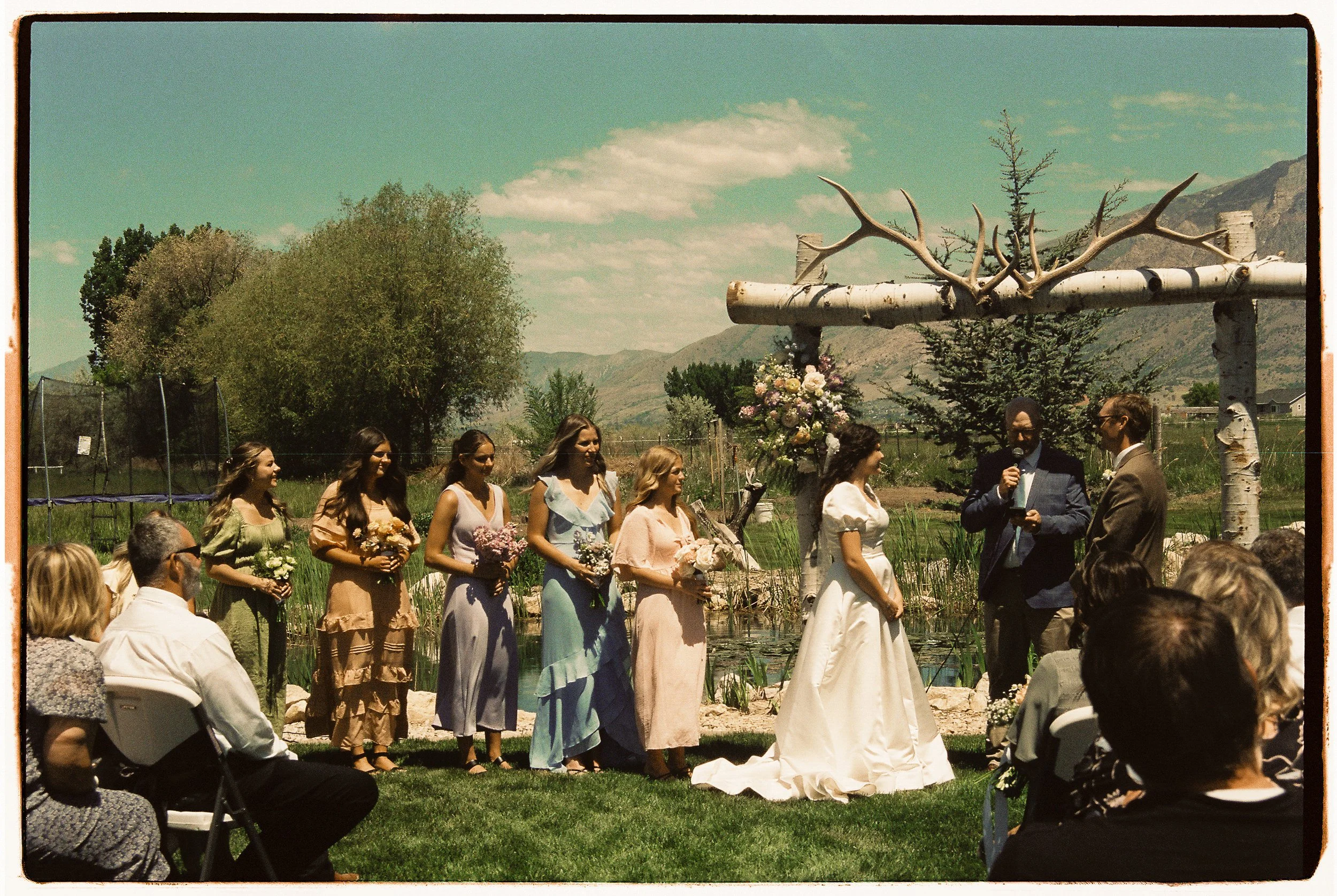 Outdoor wedding ceremony with bride and groom, bridesmaids, and officiant, under rustic arch with flowers and antler decoration, set against a mountain landscape on a sunny day.
