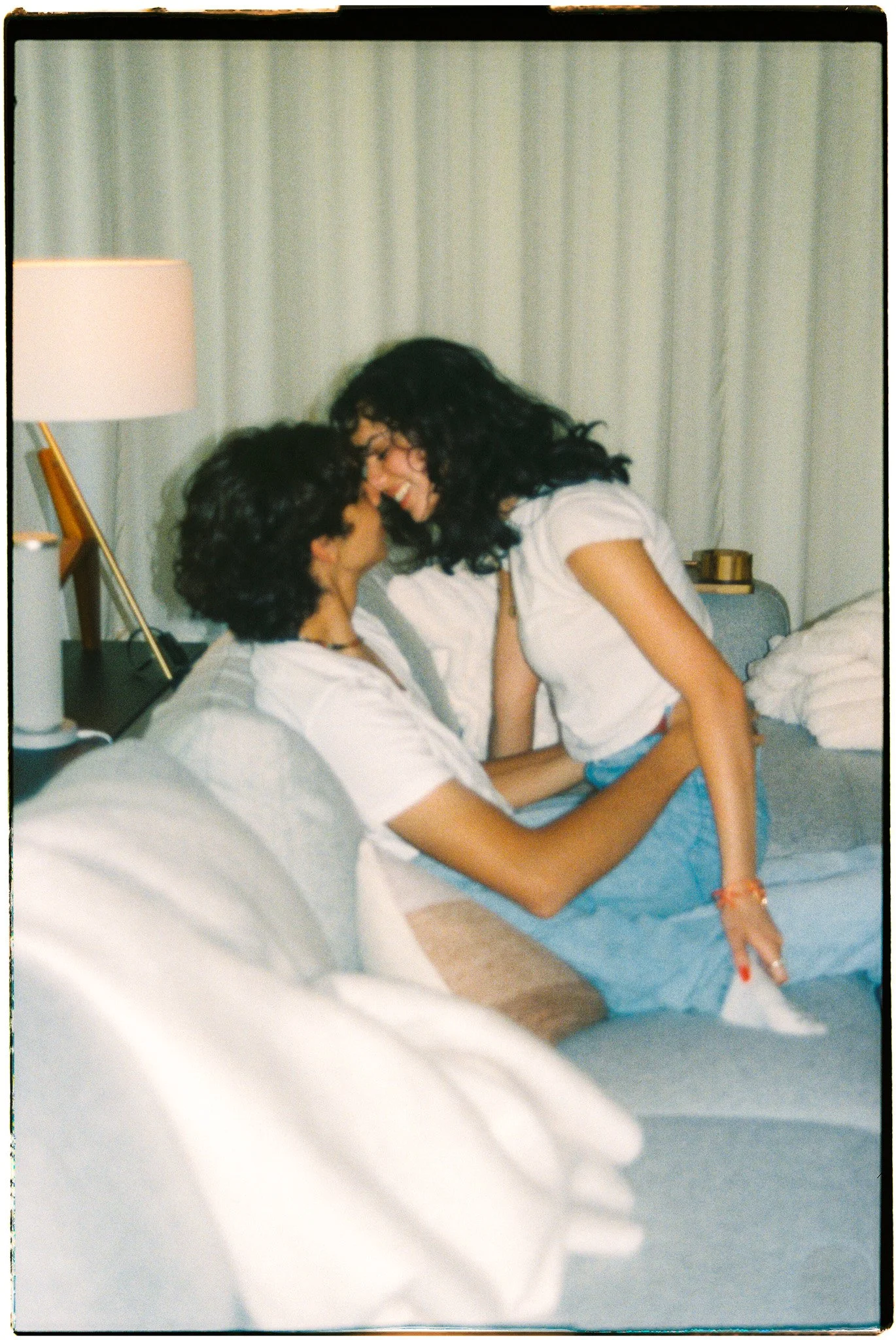 Two women with dark curly hair share a joyful moment on a bed, leaning in close and smiling, in a well-lit room with curtains and a lamp in the background.