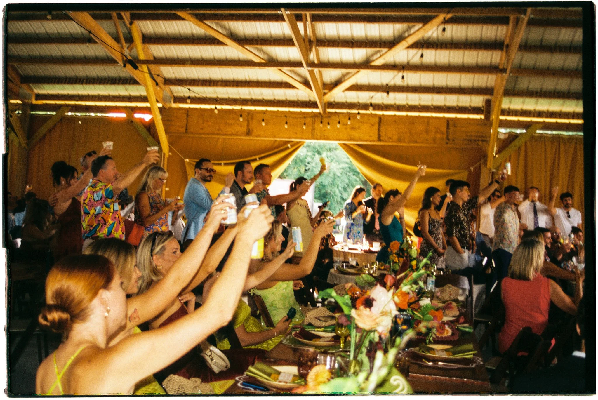 People at a celebration or party raising their glasses in a toast inside a decorated wooden venue with yellow drapes and string lights.