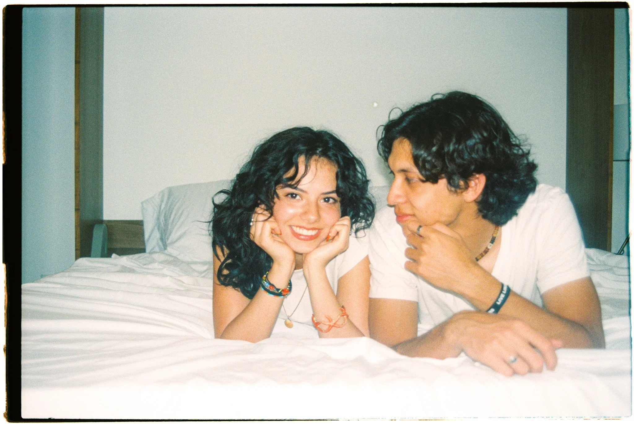 A young woman with curly black hair smiling and lying on a bed, resting her chin on her hands, with a man with curly black hair lying next to her, looking at her. Both are wearing casual white shirts and colorful bracelets.