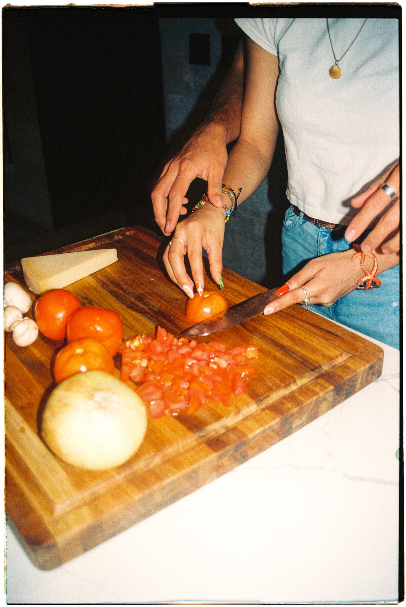 Person chopping tomatoes on a wooden cutting board with other vegetables and cheese nearby.