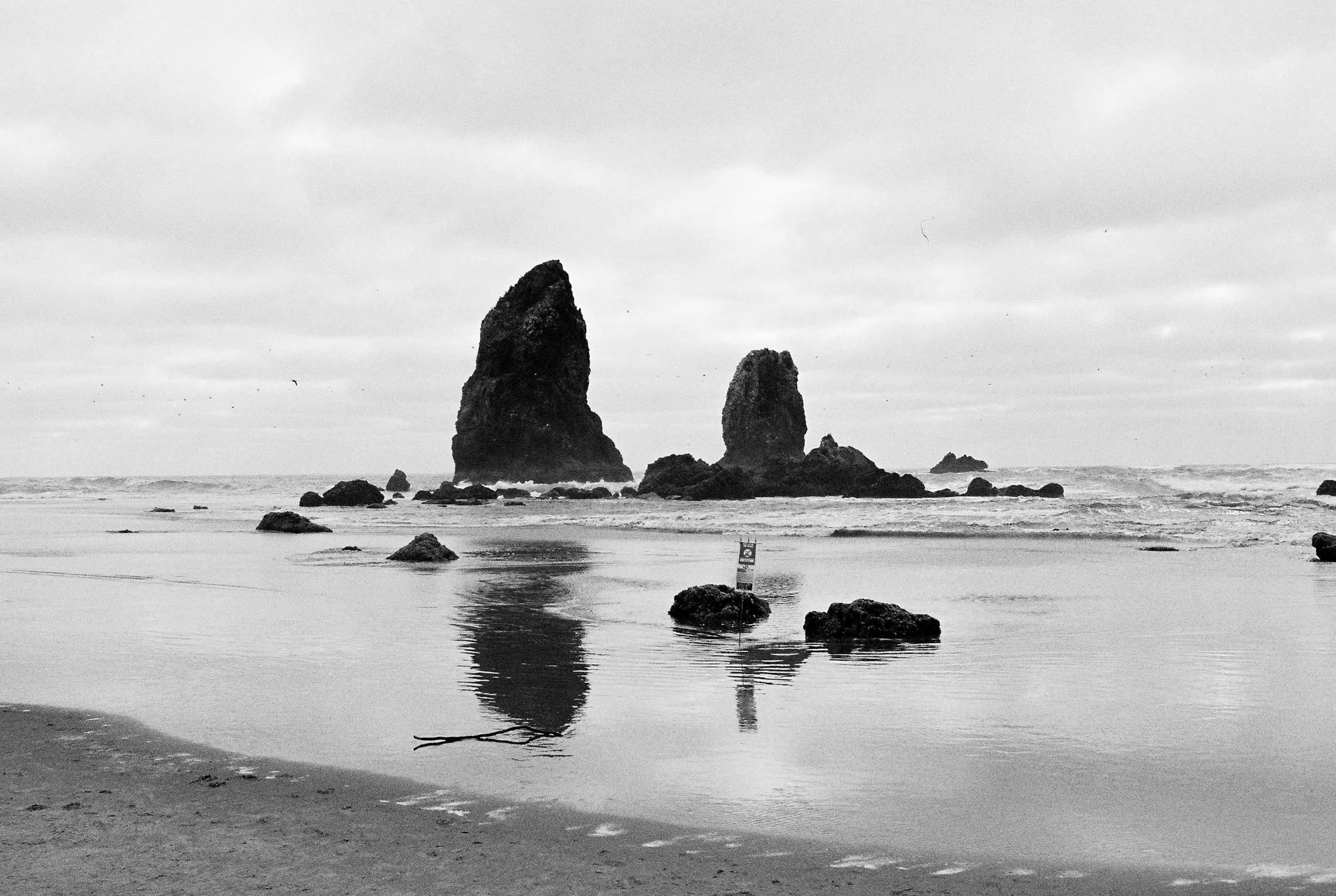 Black and white photograph of a beach with two large sea stacks in the ocean, surrounded by smaller rocks and calm water, with cloudy sky overhead.