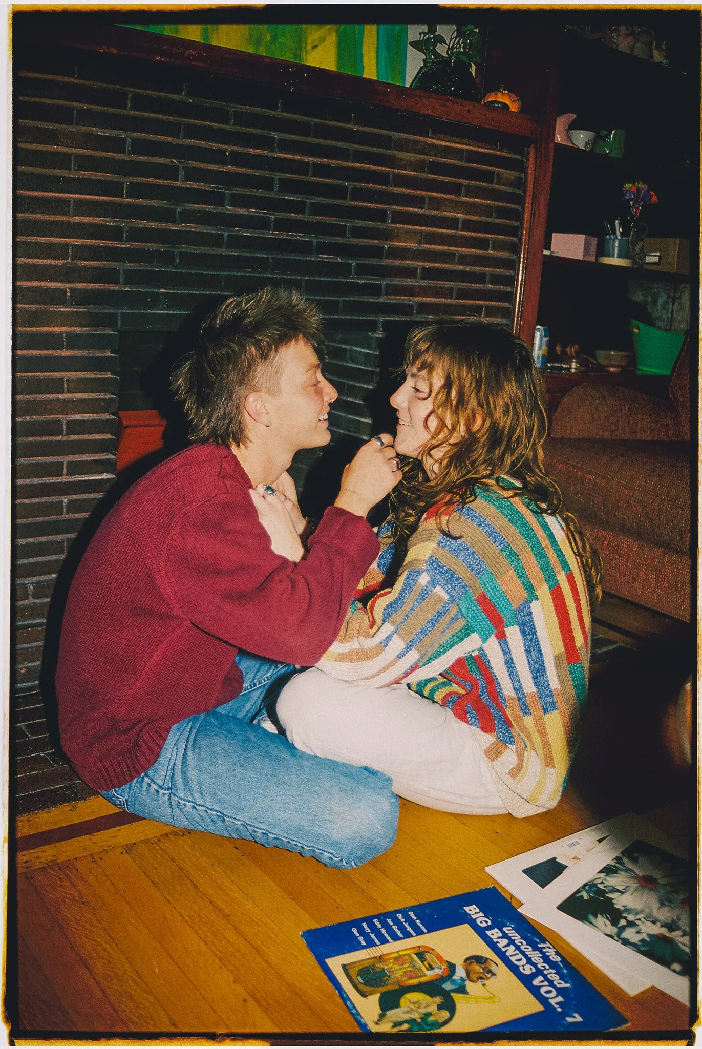 Two women sitting on the floor facing each other, smiling and holding each other's hands, with a wall, bookshelf, and magazine visible in the background.