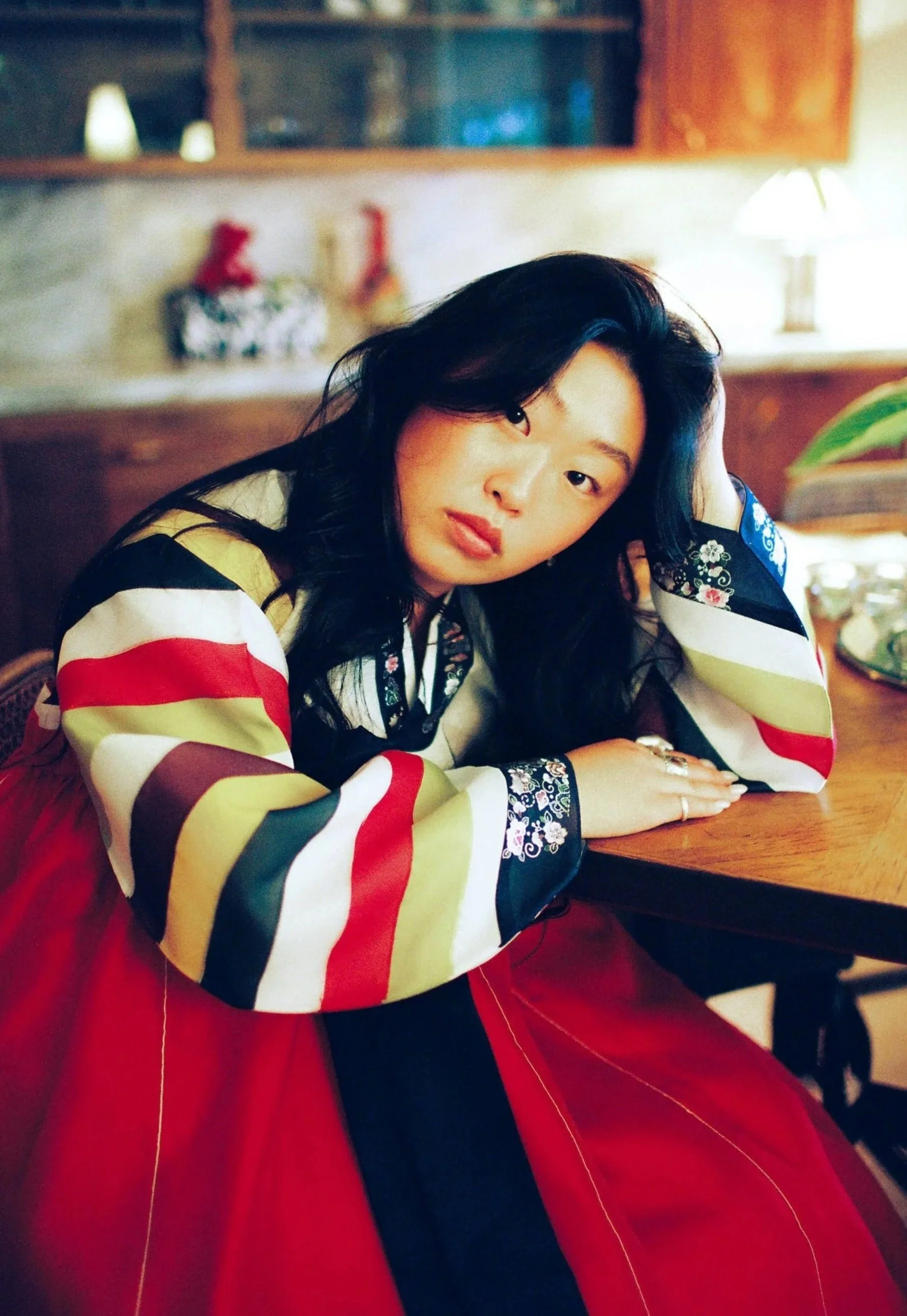 A woman sitting at a wooden table, resting her head on her left hand, with dark wavy hair, wearing a colorful top with long sleeves and a red skirt, in a cozy indoor setting with warm lighting.