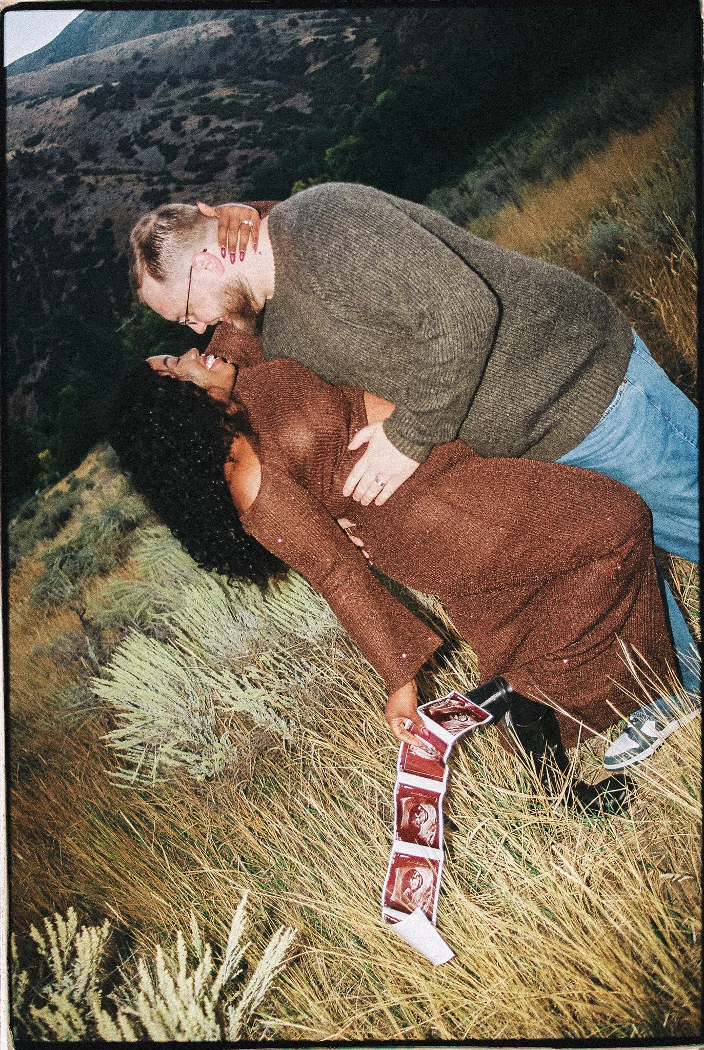 A couple passionately embrace in a field with mountains in the background. The woman is holding ultrasound images, indicating a pregnancy. The scene appears romantic and nature-inspired.