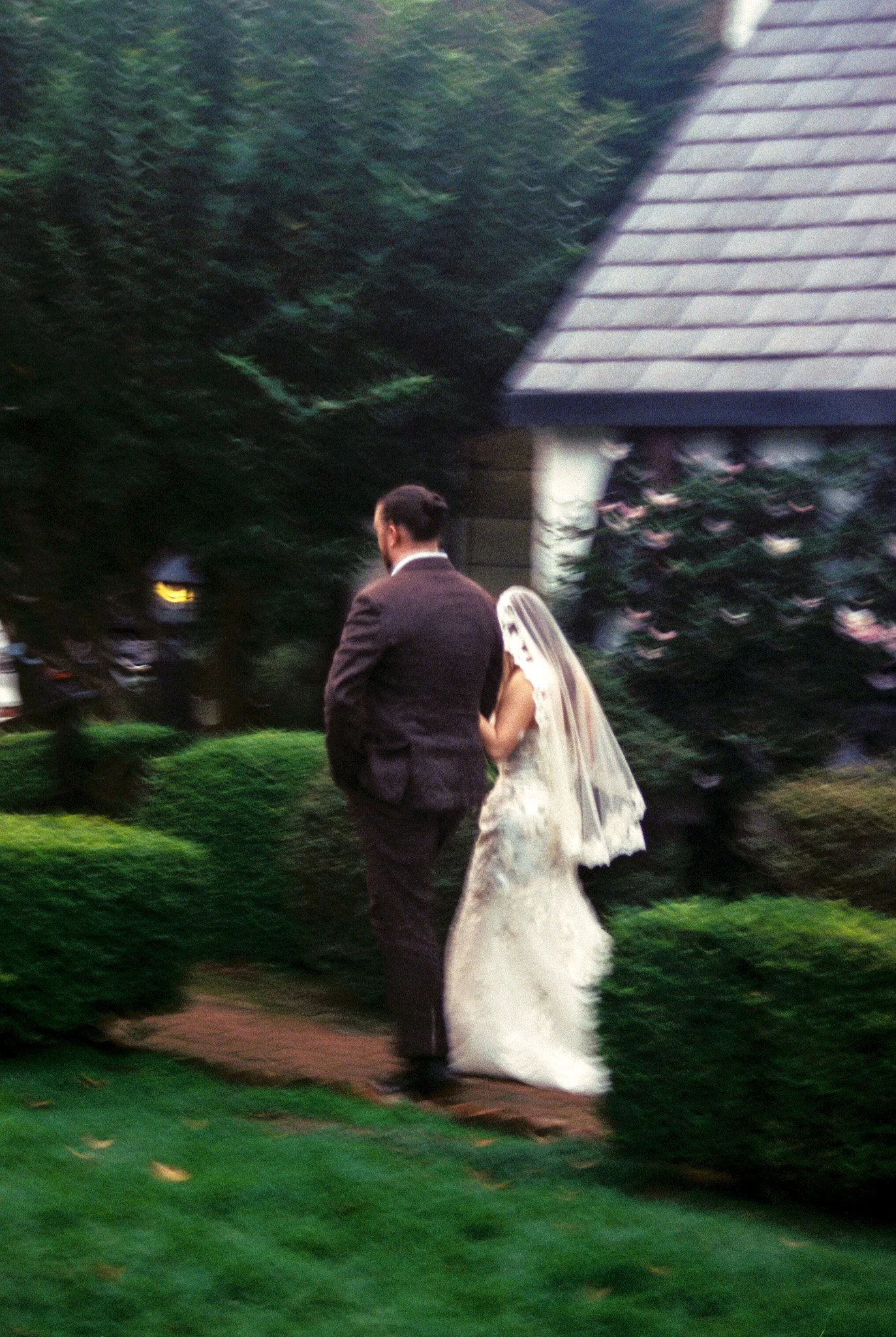 A bride in a white wedding dress with a veil walking with a groom in a dark suit on a garden pathway surrounded by greenery and bushes.