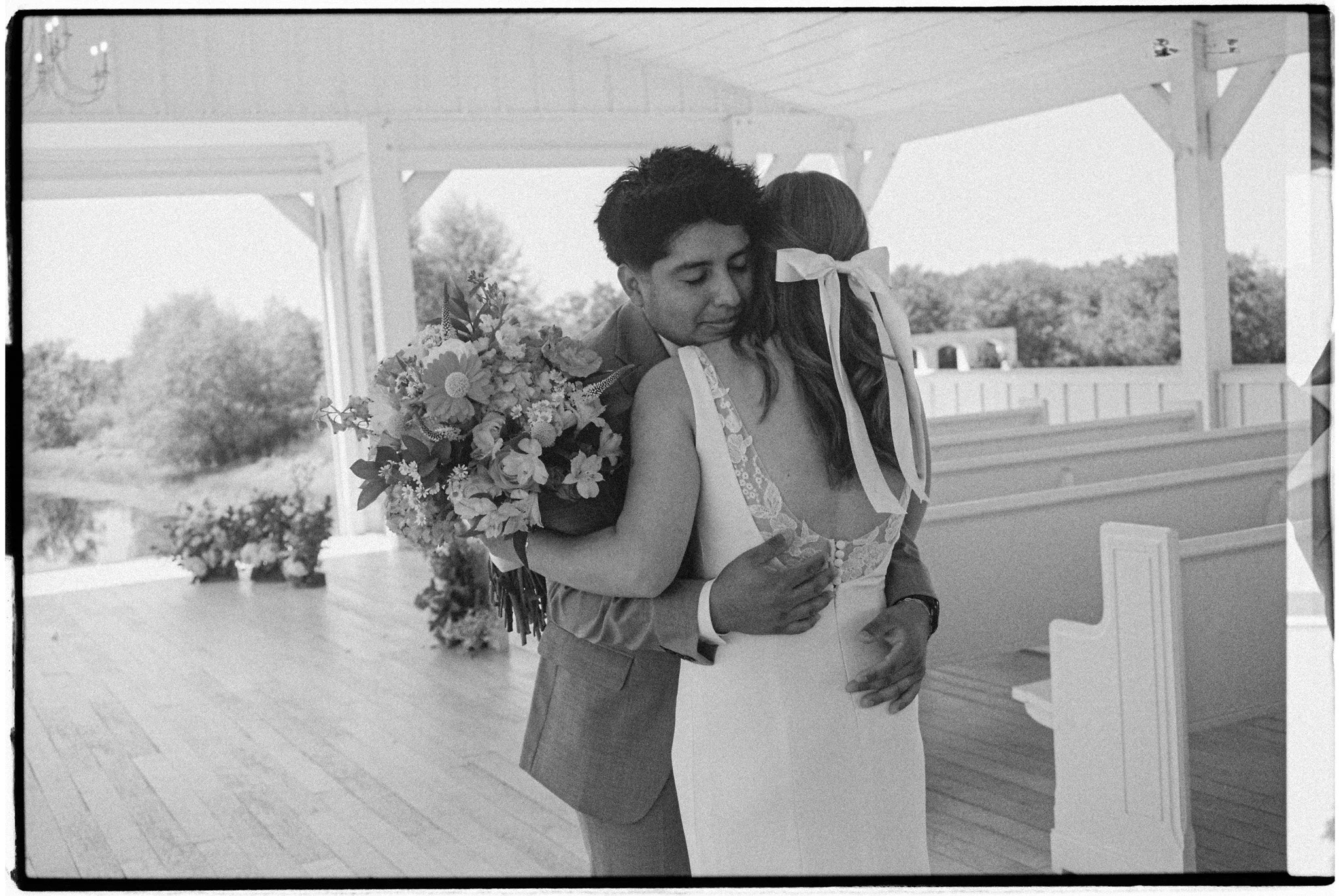 Black and white photo of a bride and groom hugging inside a pavilion with outdoor scenery in the background. The bride is holding a large bouquet of flowers, and the groom is embracing her with his eyes closed.