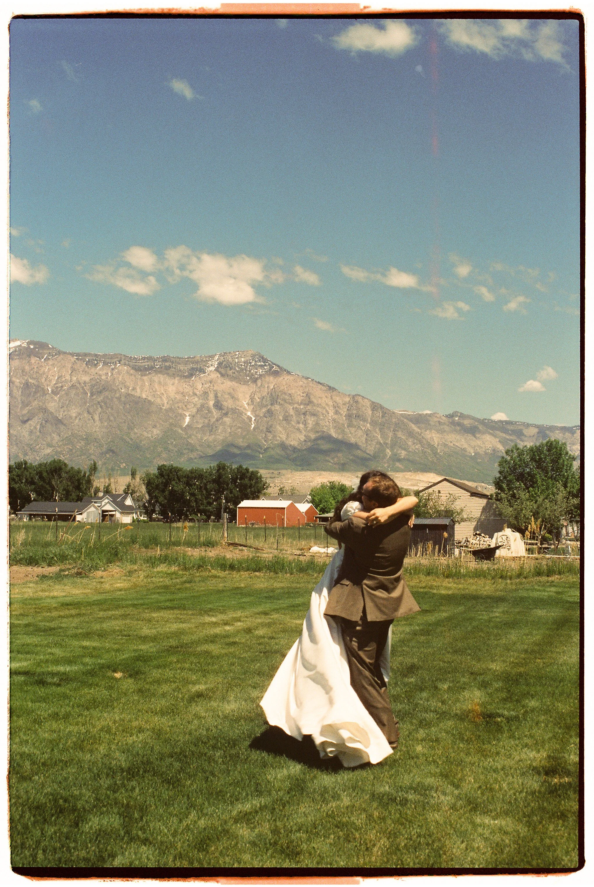 A couple in wedding attire embracing and dancing on a green lawn with mountains, trees, and houses in the background under a partly cloudy sky.