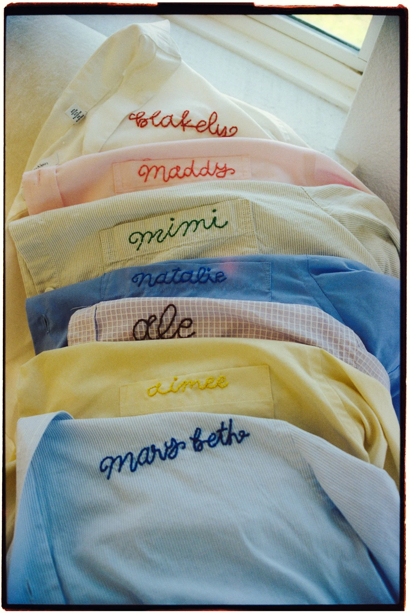 A row of colorful embroidered shirts, each with a different name, laid flat on a surface near a window.