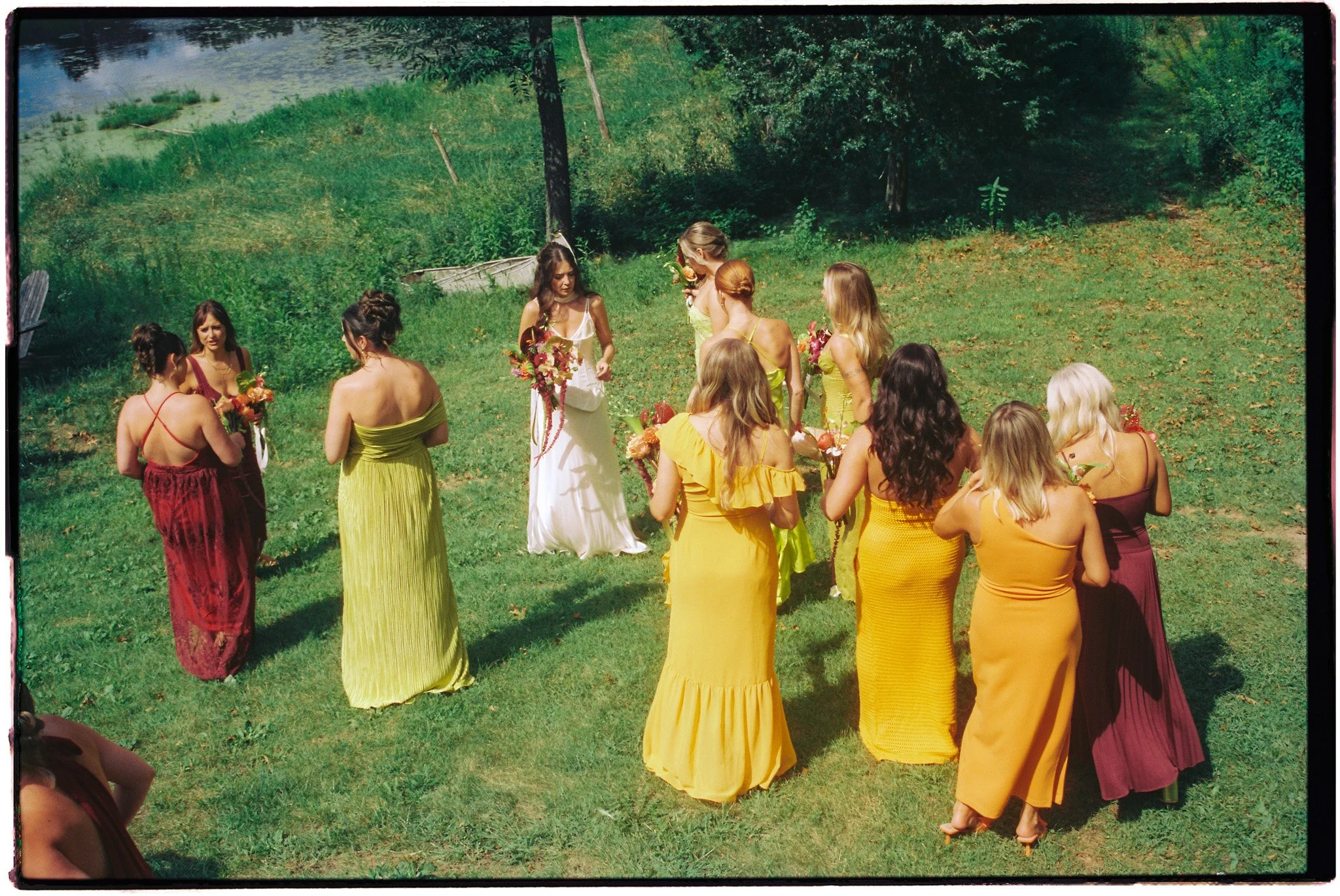 A wedding ceremony outdoors with a bride and bridesmaids in colorful dresses holding bouquets, gathered on a grassy area near a lake surrounded by trees.