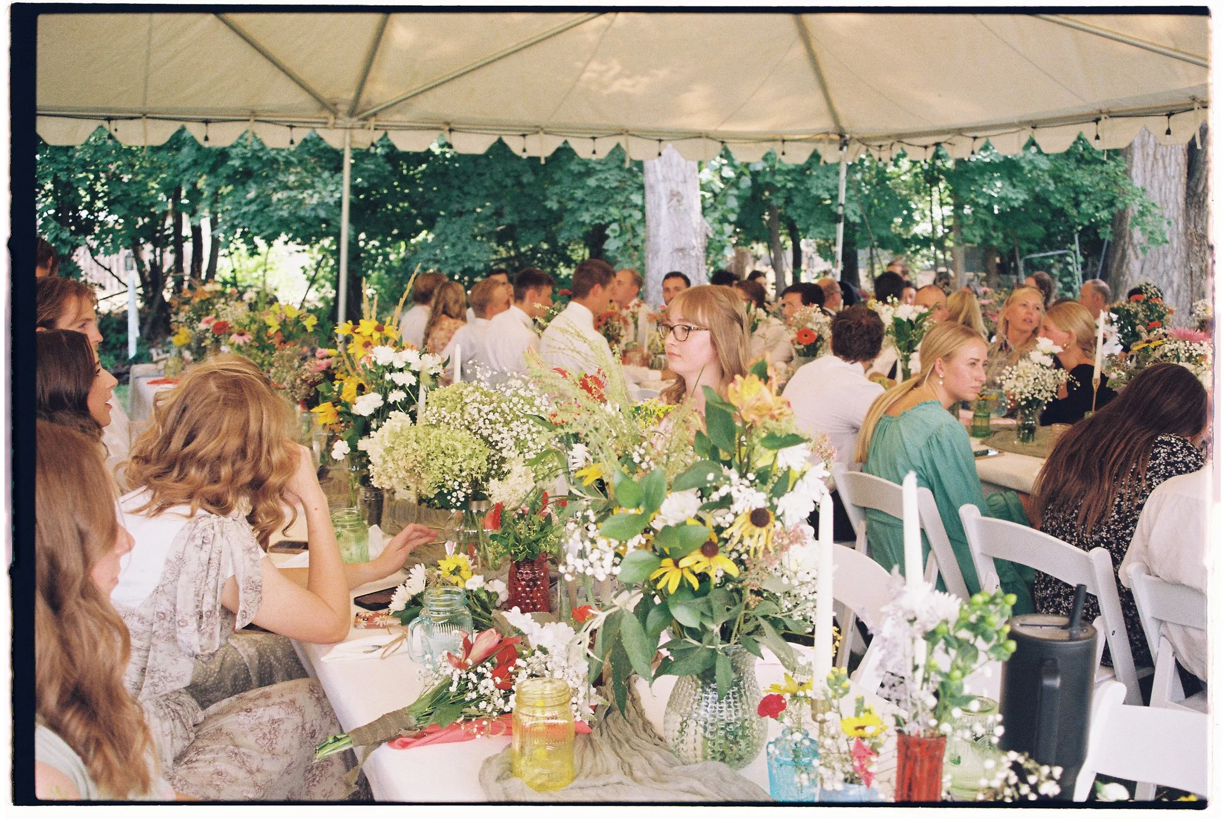 People seated at long tables under a canopy, decorated with large floral arrangements, during an outdoor gathering surrounded by trees.