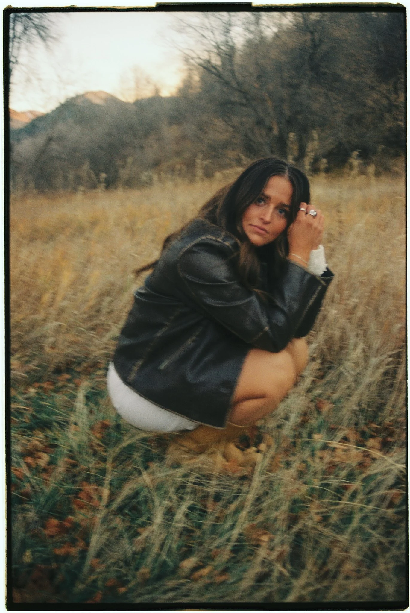 A woman squatting in a field of tall grass and dried leaves, wearing a black leather jacket, white shorts, and tan boots. She has long dark hair and is looking at the camera with a neutral expression, with hills and trees blurred in the background.