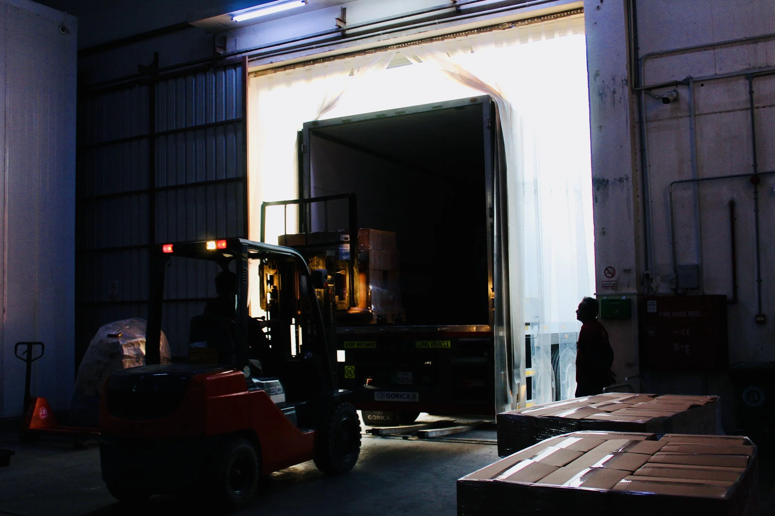 Inside a warehouse, a forklift is moving goods into a truck. There are boxes on the ground and a person standing near the wall.