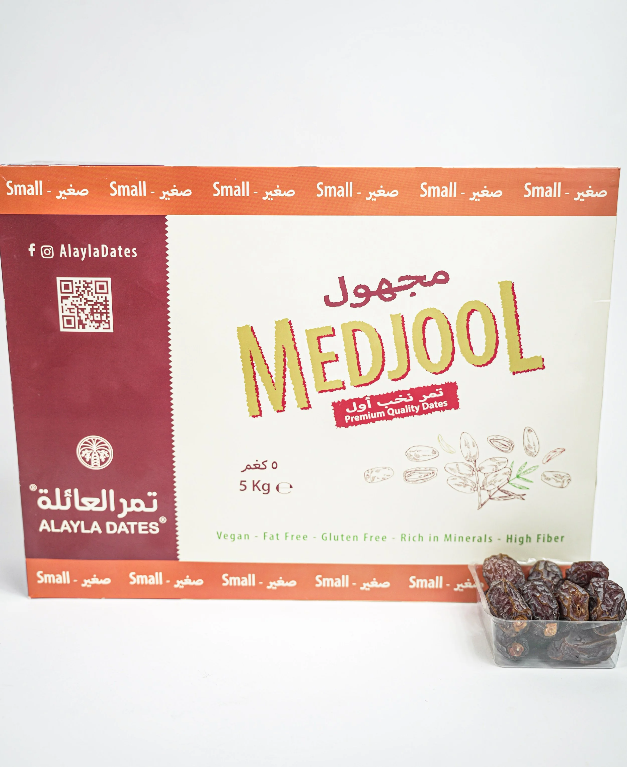 A box of Medjool dates with some dates in a small clear plastic container in front of it. The box has text in both Arabic and English and displays nutritional benefits.