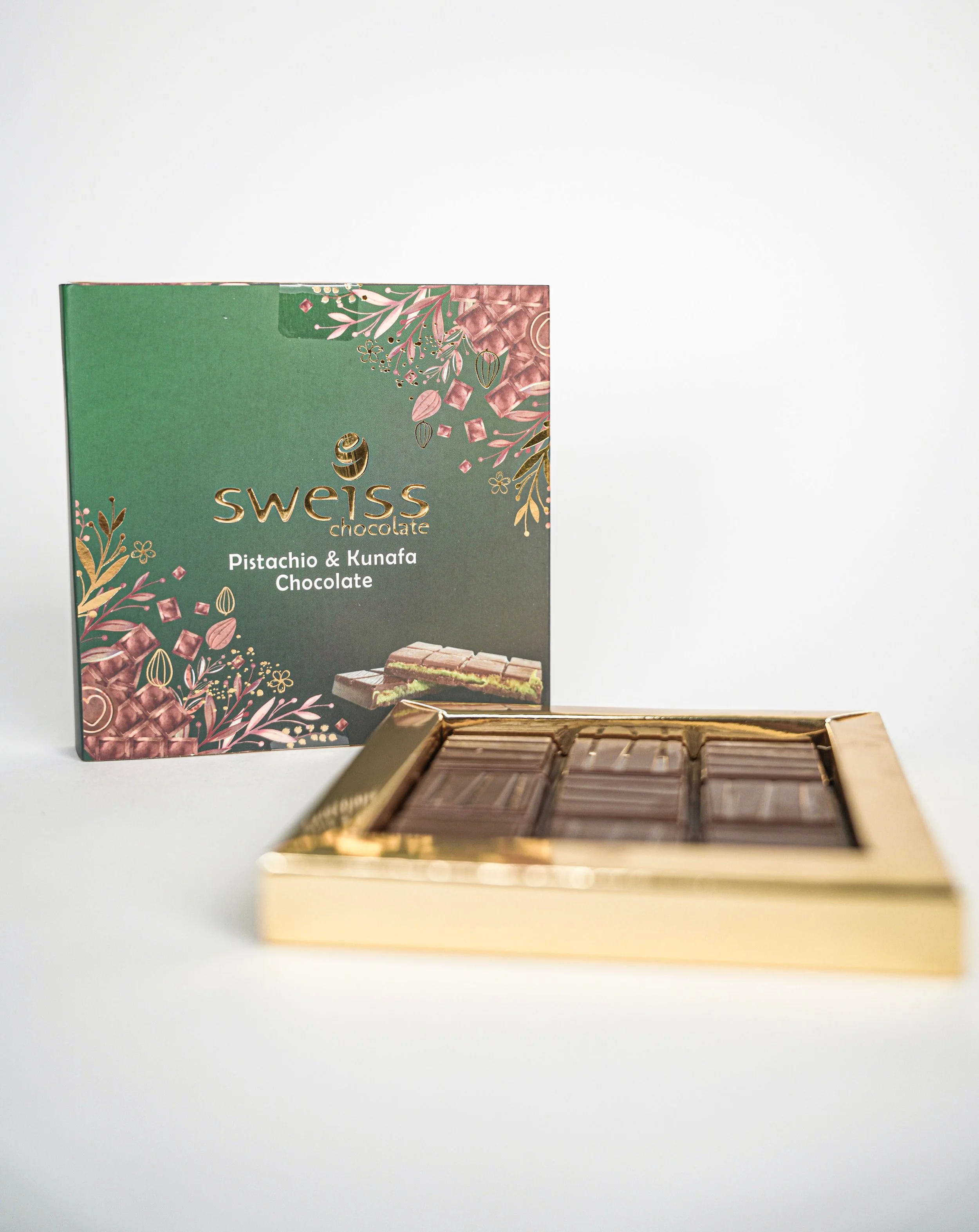 A box of Swiss chocolate with pistachio and kunafa flavor, partially opened showing four chocolate bars inside, and decorative packaging with floral motifs.