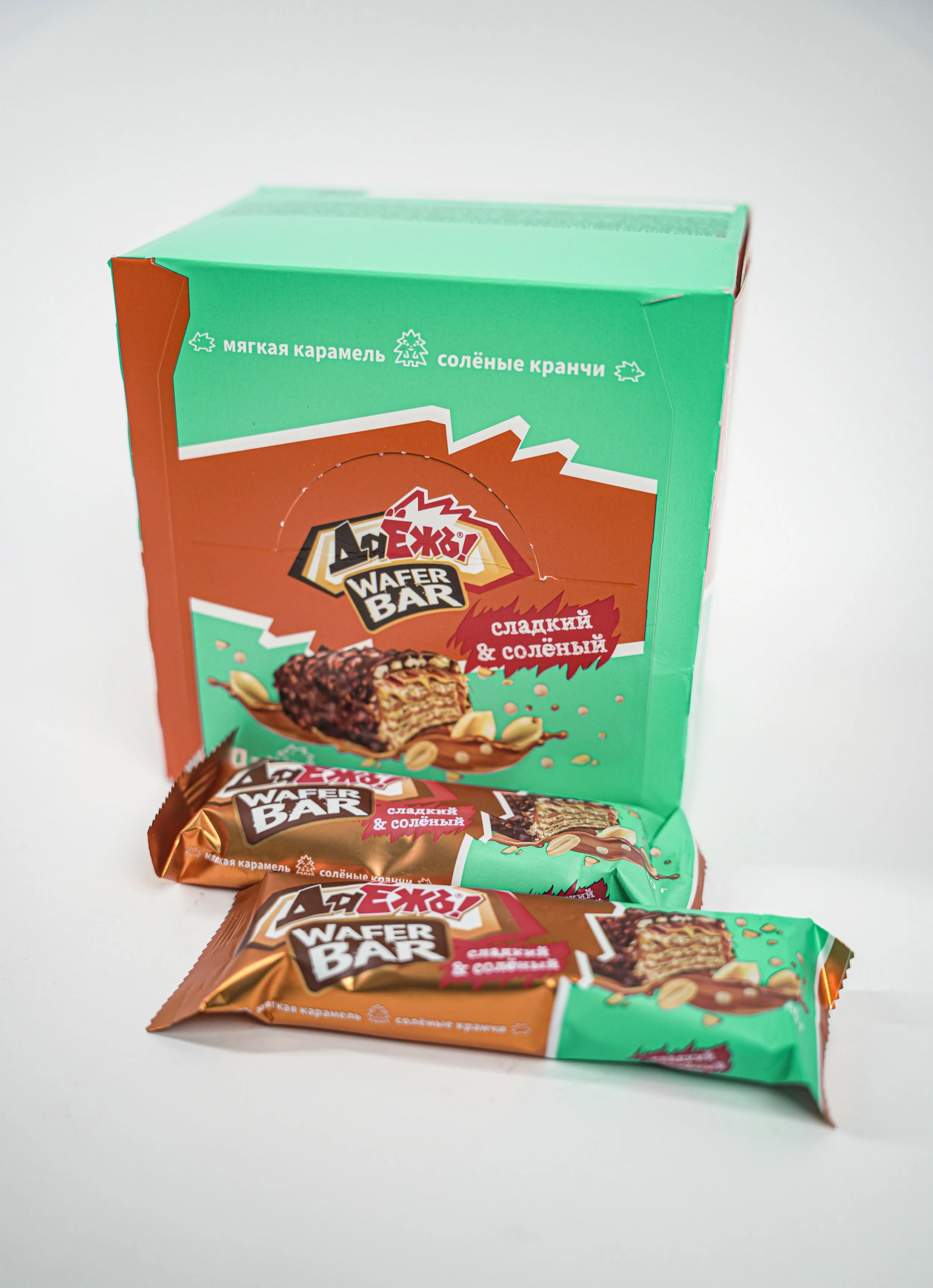 A green and orange box of wafer bars with two wrapped wafer bars in front of it. The packaging has an image of a wafer with caramel and chocolate, and some almonds, with Russian text indicating the flavor as sweet and salty.