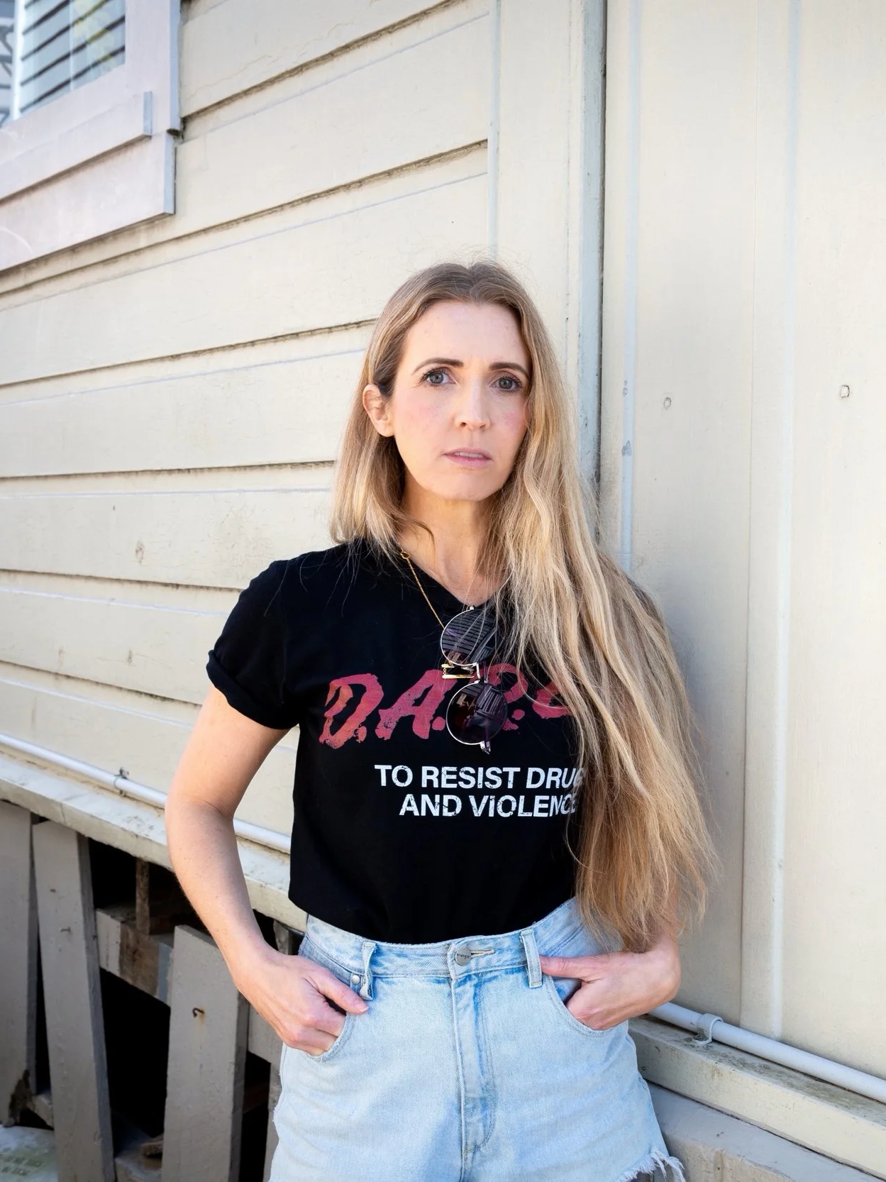 A woman with long blonde hair standing outdoors against a beige wall, wearing a black T-shirt with red and white text and high-waisted light blue denim shorts, with sunglasses hanging from her shirt.
