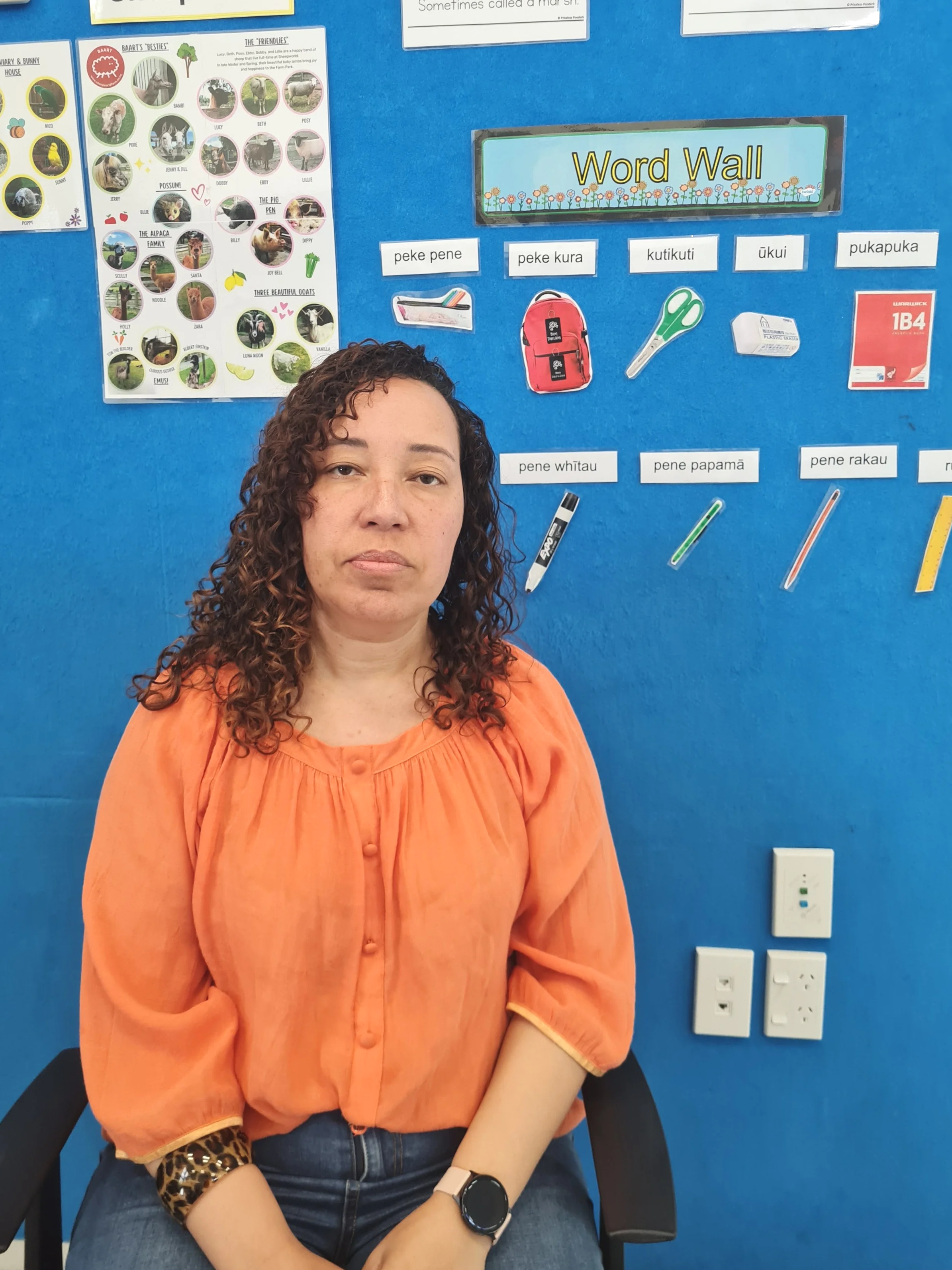 A woman with curly hair wearing an orange blouse sitting in a classroom with a blue wall behind her, which has educational posters and labels for words and school supplies.