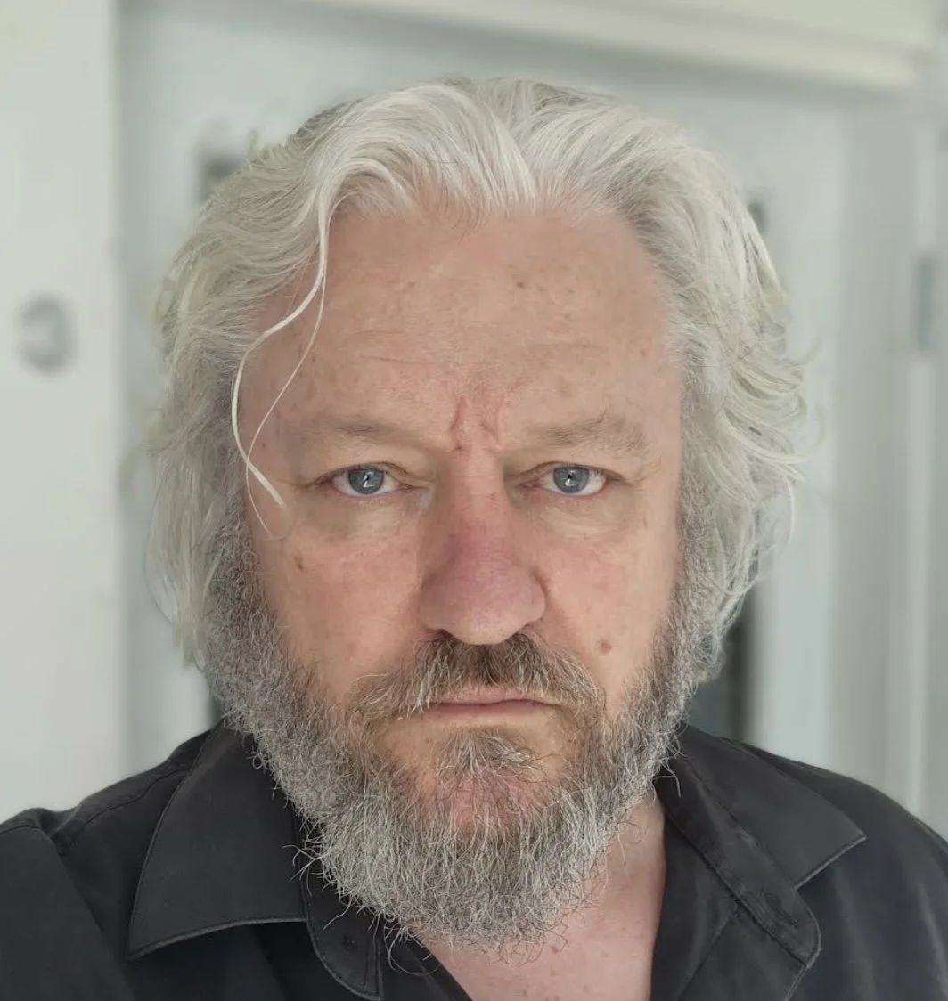 Close-up of a middle-aged man with long gray hair, blue eyes, and a beard, wearing a black shirt, standing indoors.