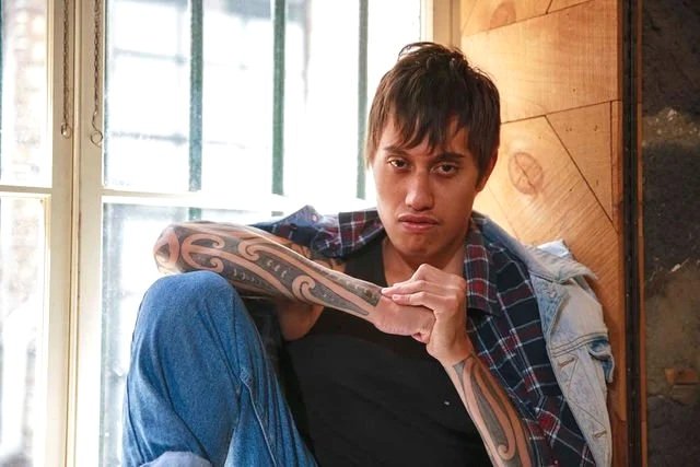 Young man with short brown hair and tattoos on his arms, wearing a black shirt, a denim jacket, and plaid shirt, sitting with one knee up, inside a wooden room near a window.