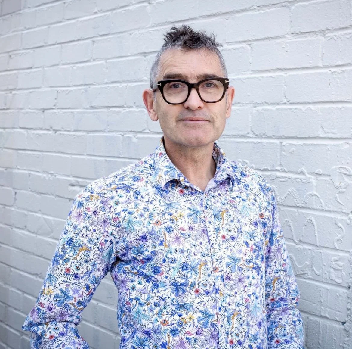 A man with glasses and short gray hair wearing a colorful floral shirt, standing against a white brick wall.