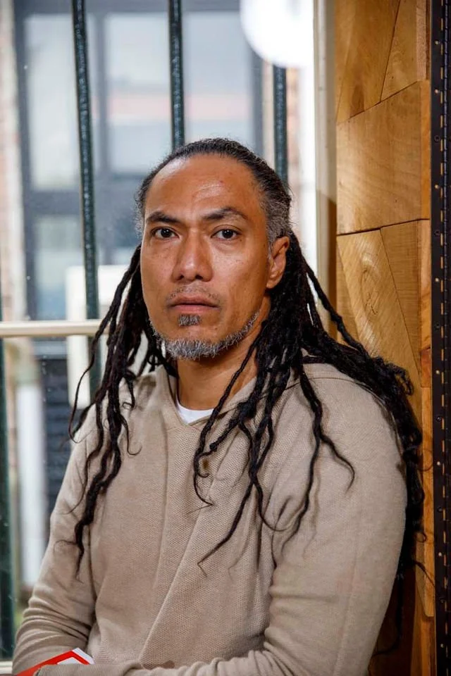 A man with long dreadlocks, facial hair, and a serious expression, sitting indoors near a window with a wooden wall behind him.