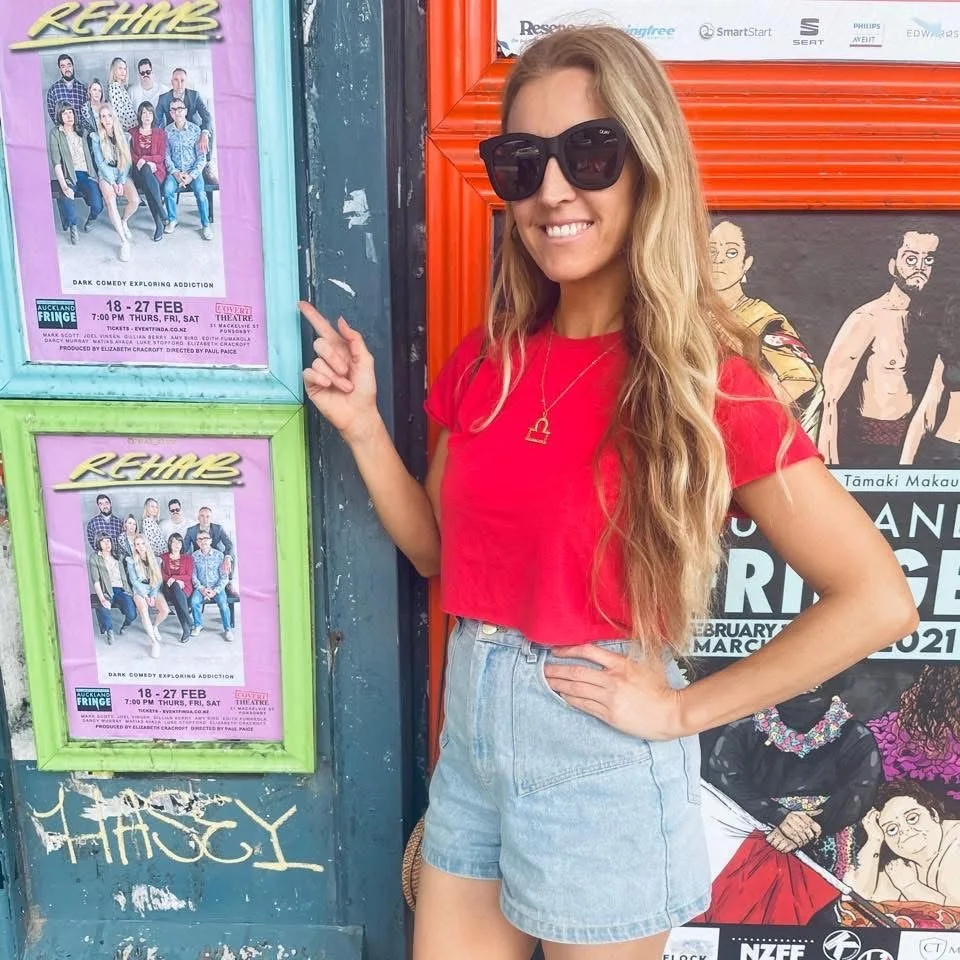 A woman with long, blonde hair, wearing a red shirt, denim shorts, large black sunglasses, and a gold necklace, stands outdoors next to Auckland Fridge Festival 2021 posters. She is smiling and pointing at two framed A4 REHAB posters.