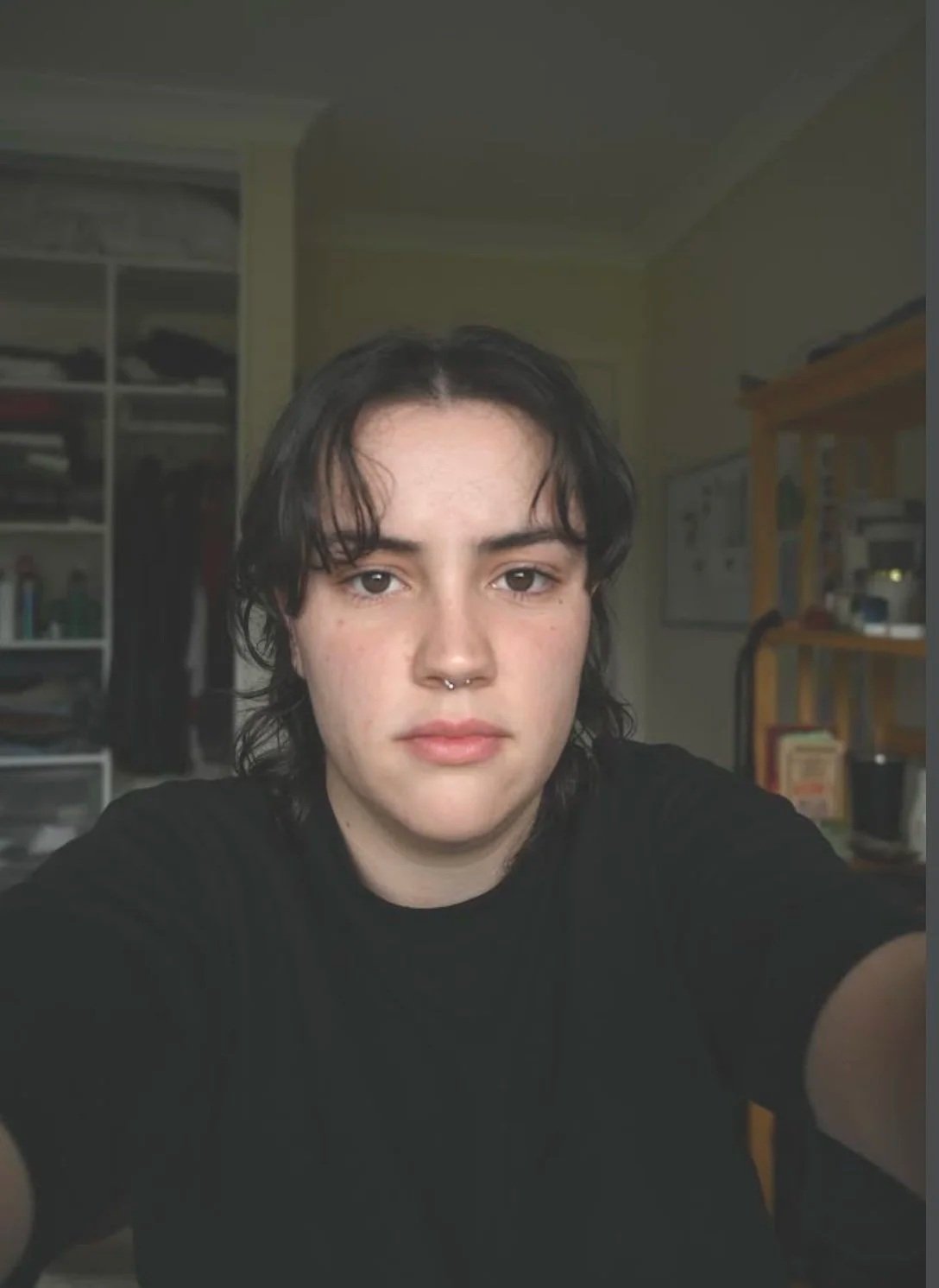 A person with short dark hair, light skin, and a septum piercing looking into the camera, with a room of shelves and books in the background.