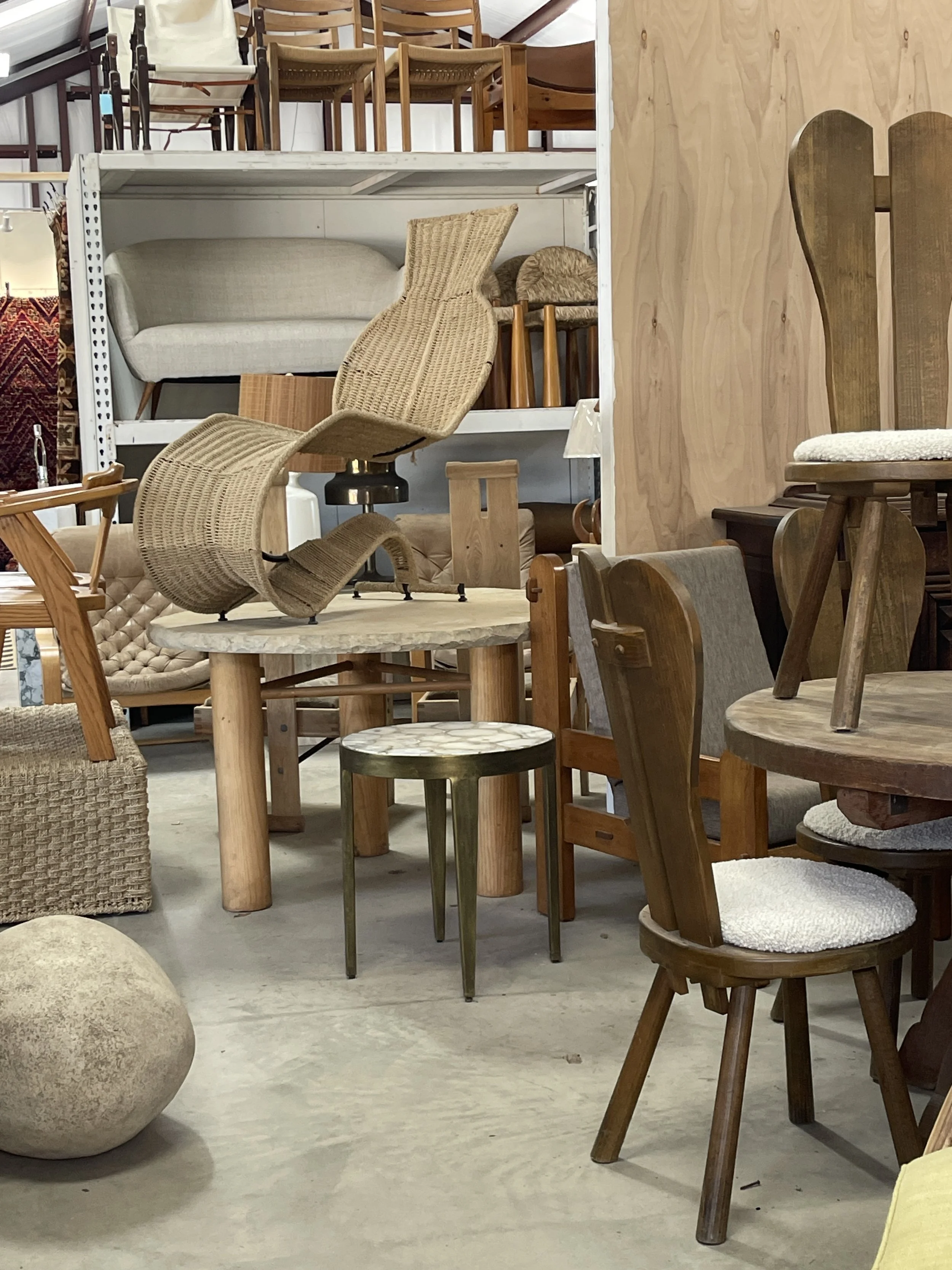 Store display of various wooden and fabric chairs and stools, with a beige upholstered sofa, some stacked furniture, and a large round beige ottoman in the foreground.