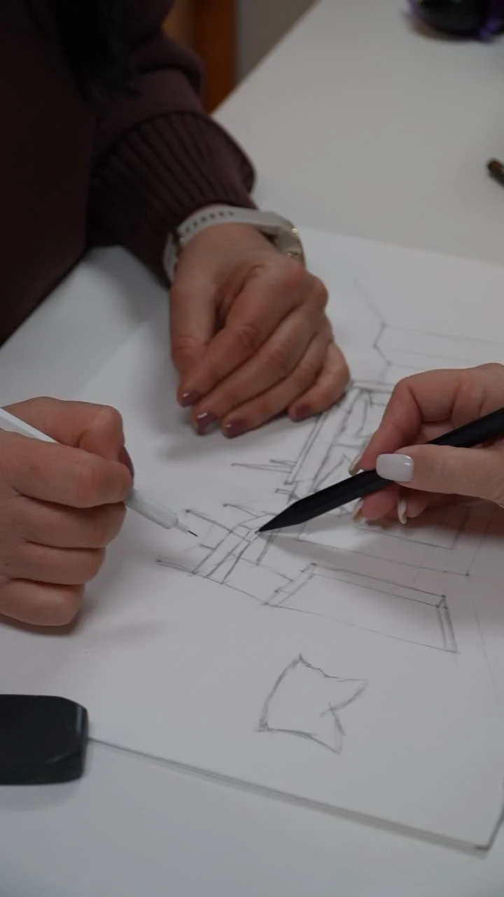 Two people working on an architectural drawing or sketch on white paper with a pencil and pen.