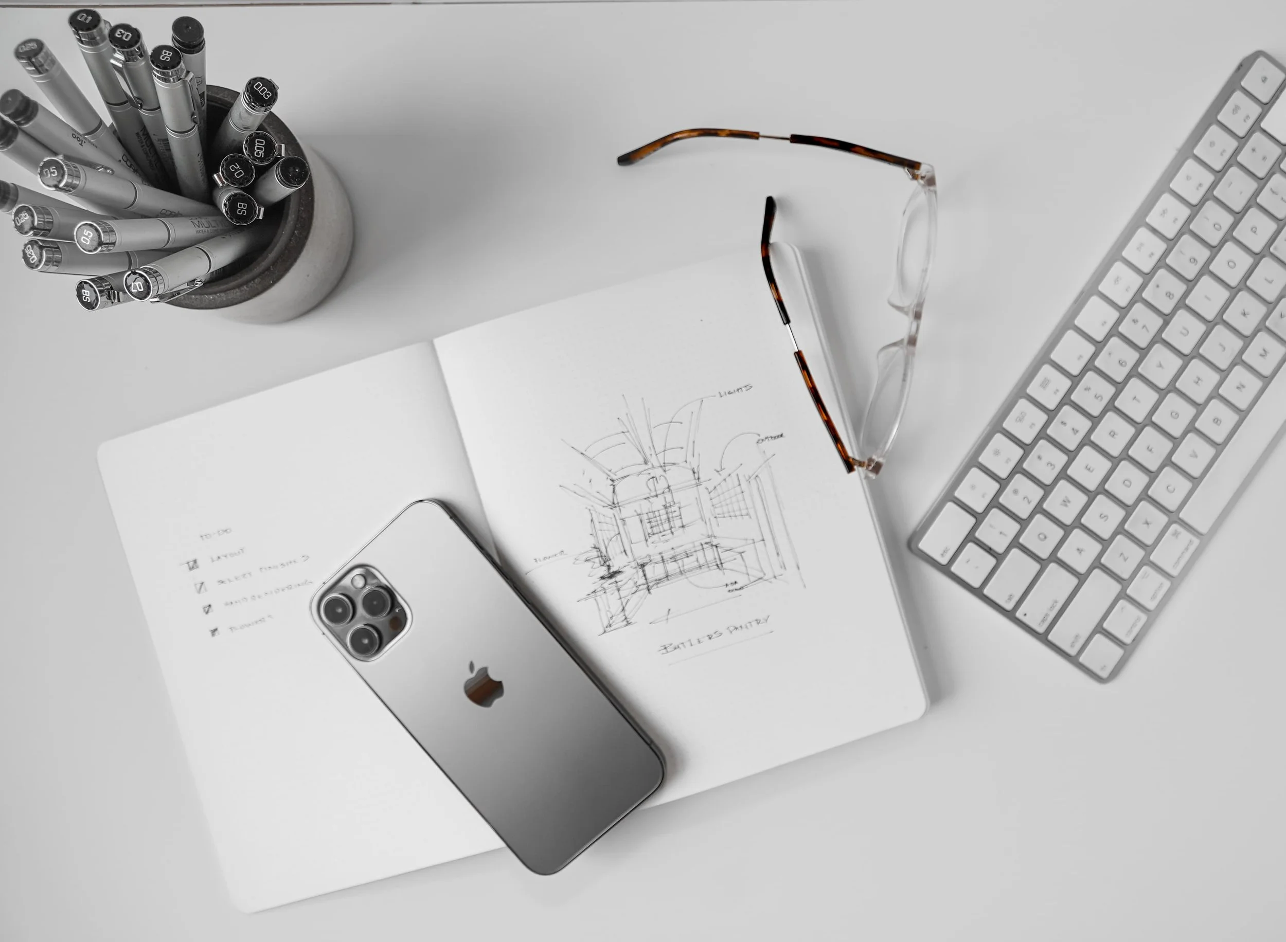 A white desk featuring a silver iPhone, a wireless keyboard, a pair of tortoise-shell glasses, an open sketchbook with architectural drawings, a sheet with a to-do list, a notebook, and a container of black and gray pens.
