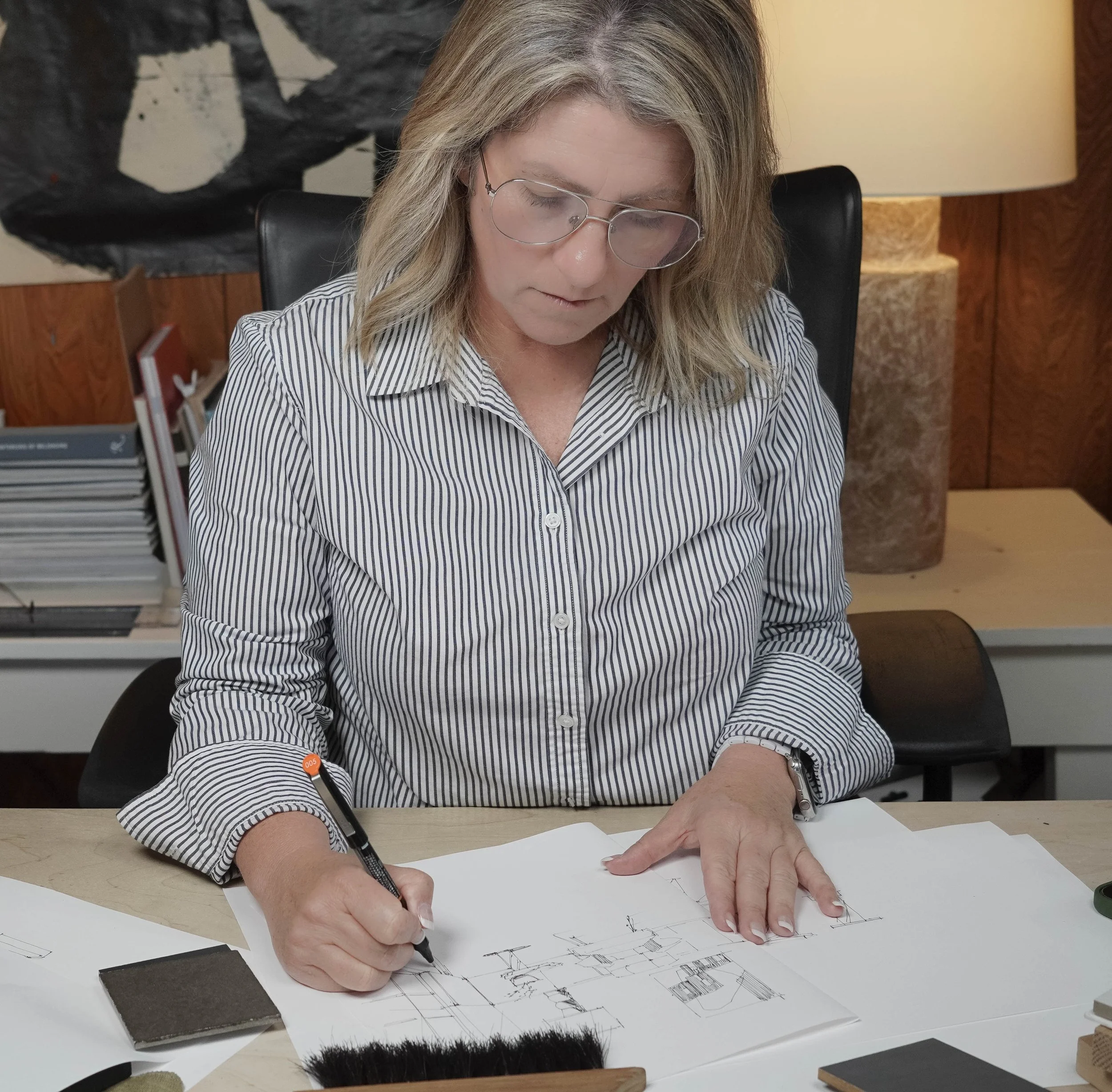 A woman with blonde hair and glasses wearing a striped shirt is sitting at a desk, drawing architectural sketches on paper with a black pen. There are color samples and design tools on the desk, and a lamp and bookshelf are in the background.