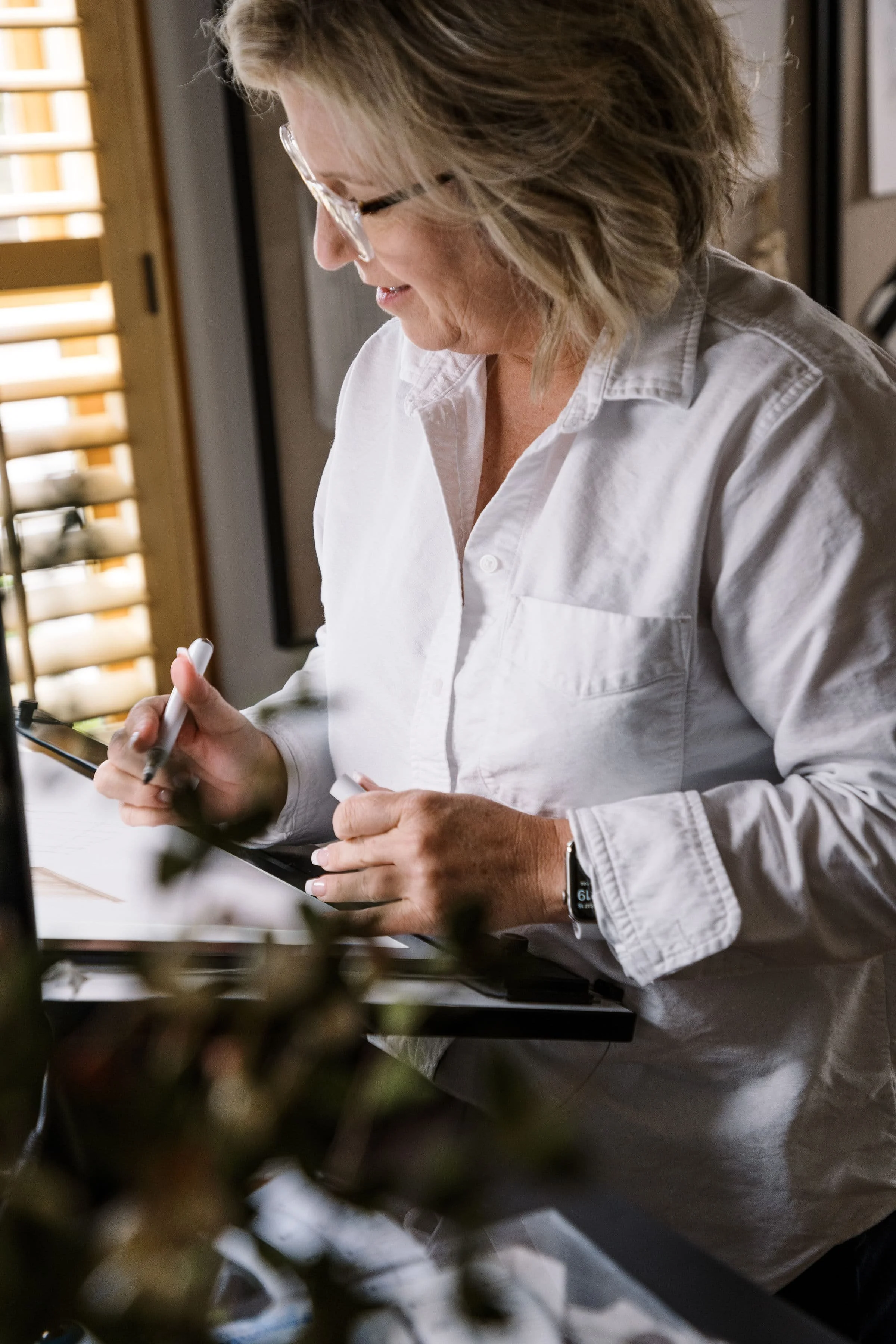 Woman with glasses writing on a tablet in a well-lit room.